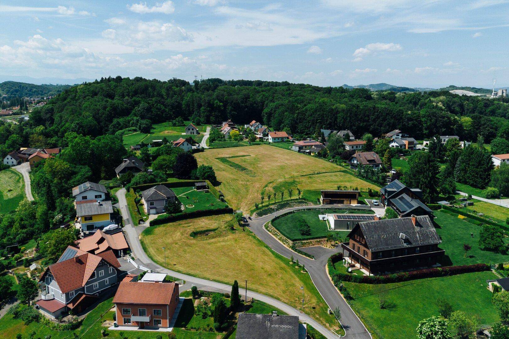 Weitläufige Luftaufnahme einer Wohnsiedlung eingebettet in eine grüne Hügellandschaft unter blauem Himmel.