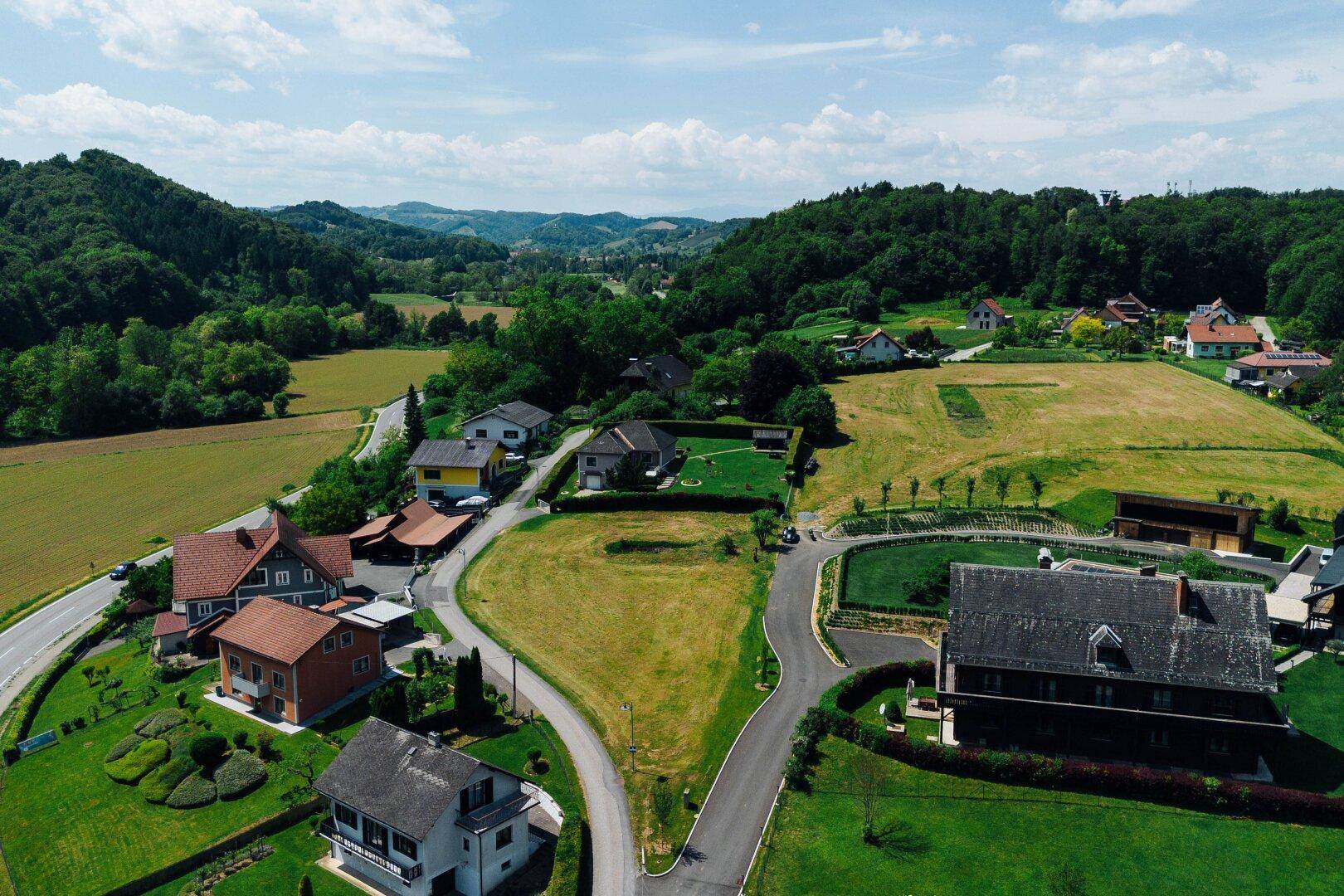 Luftbild einer Wohngegend mit Straßen, Häusern und weiten grünen Feldern in einer hügeligen Landschaft.