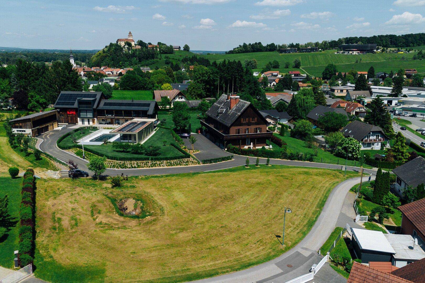 Luftaufnahme einer Wohnsiedlung mit einem unbebauten Grundstück, modernen Häusern und einem Schloss im Hintergrund.