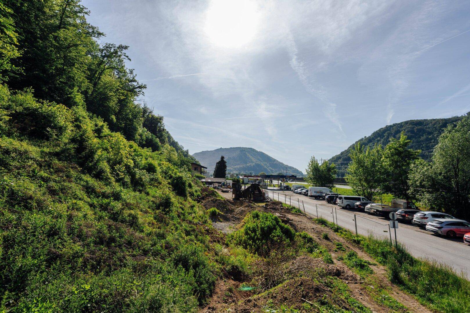 Eine Aufnahme der Umgebung mit einer Straße, grünen Hügeln und Bergen im Hintergrund unter hellem Himmel.