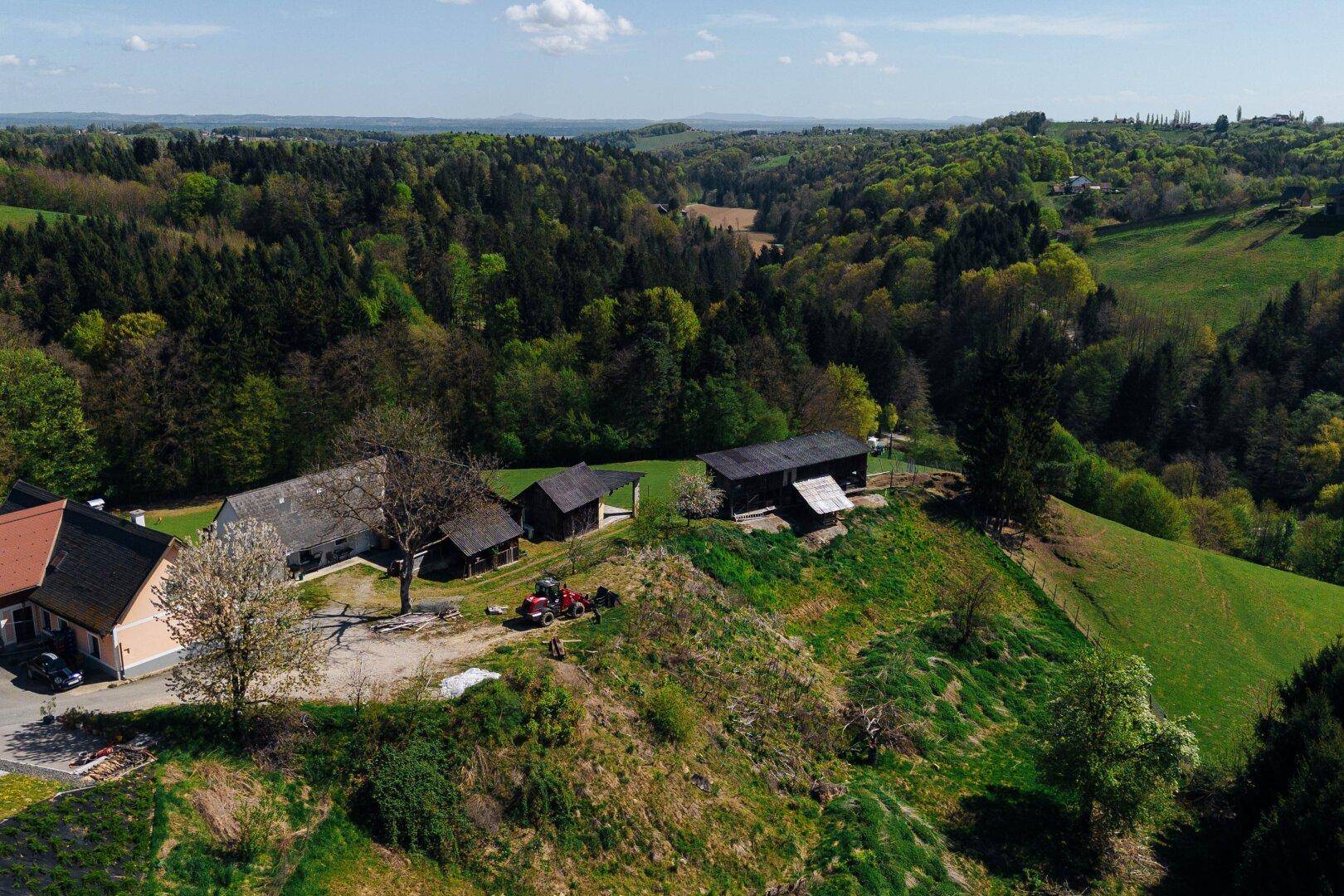 Panoramablick auf ein ländliches Anwesen mit mehreren Gebäuden und einem Traktor inmitten grüner Hügel.