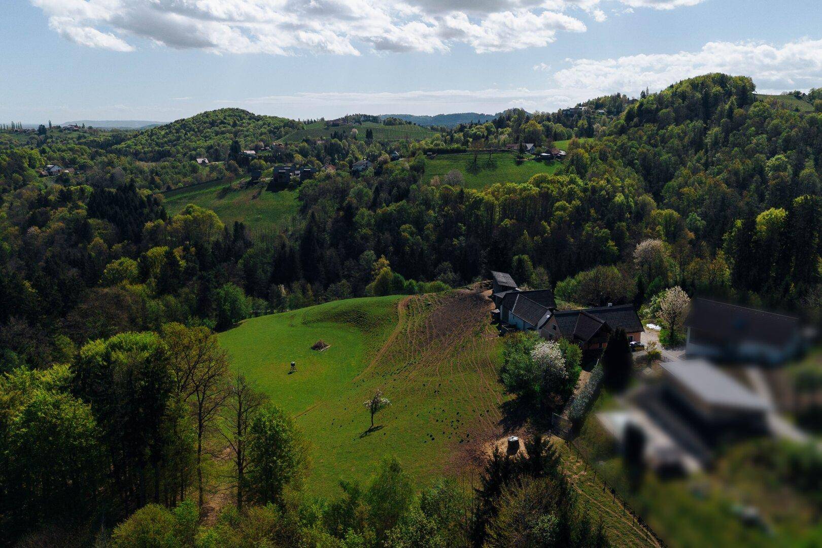 Panoramablick auf ein Anwesen in einer idyllischen grünen Hügellandschaft unter blauem Himmel.