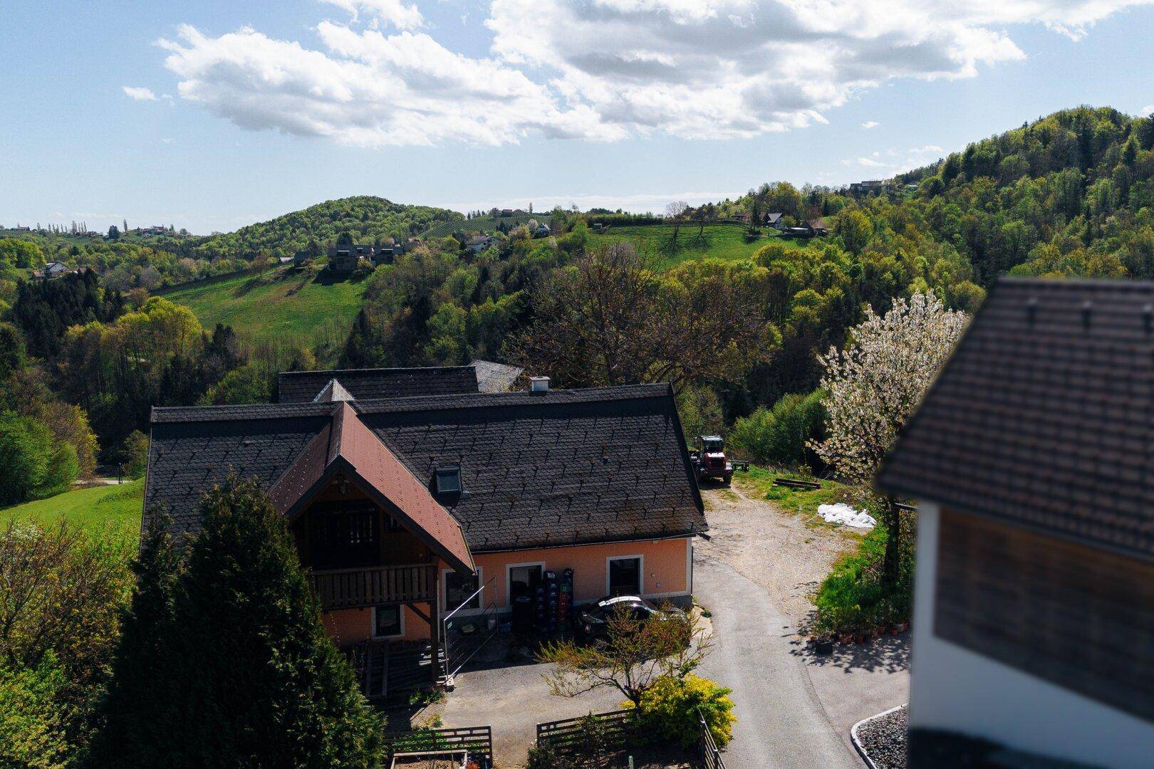Ländliches Haus mit rotem Dach und Zufahrt, umgeben von grünen Hügeln und einem Traktor im Hintergrund.
