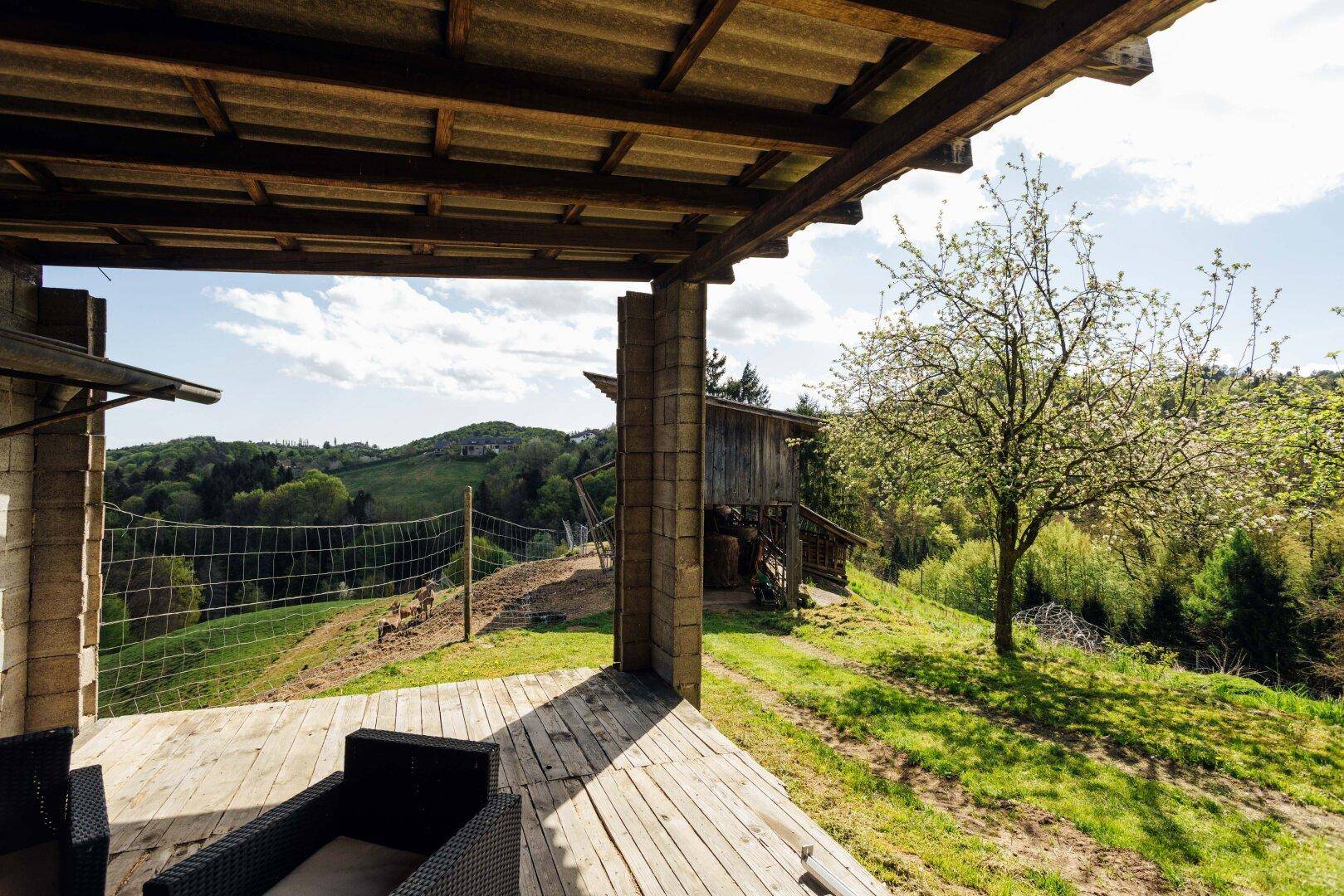 Gemütlicher Sitzbereich auf einer Holzterrasse mit weitem Blick über ein grünes Tal und eine Scheune.