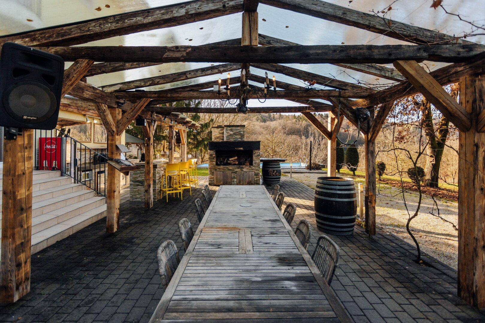 Rustikaler Essbereich im Freien unter einer Pergola mit großem Holztisch und Außenkamin.