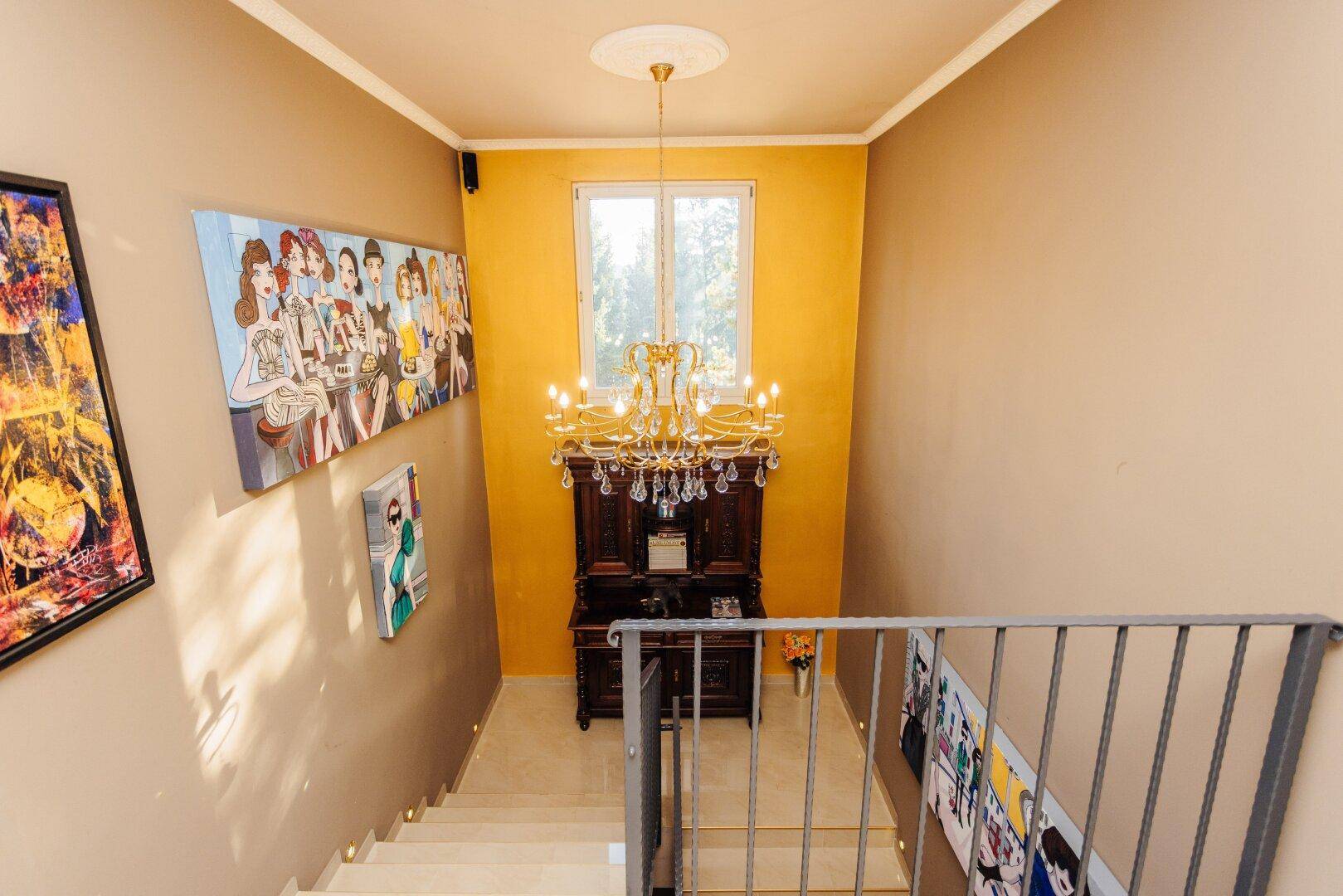 Blick von oben auf die Treppe mit einem prunkvollen Kronleuchter und farbenfrohen Bildern.