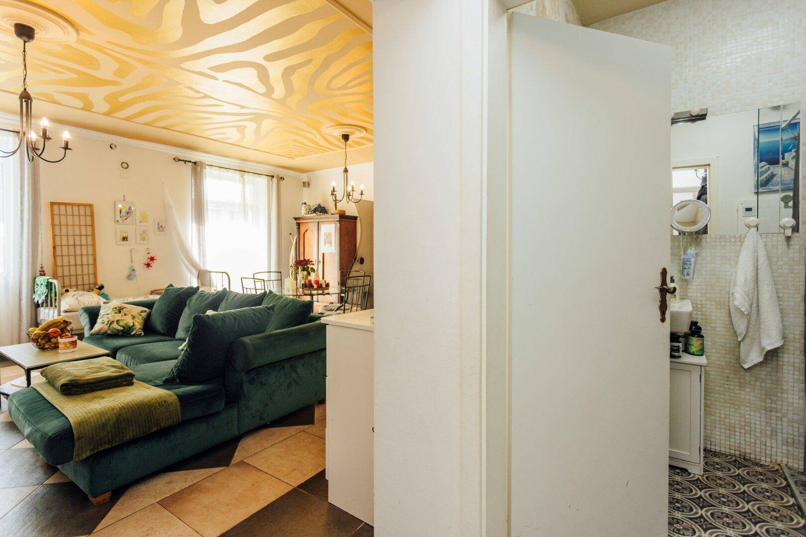 Wohnzimmer mit grünem Sofa, gemusterter Decke und Blick in ein angrenzendes Badezimmer.