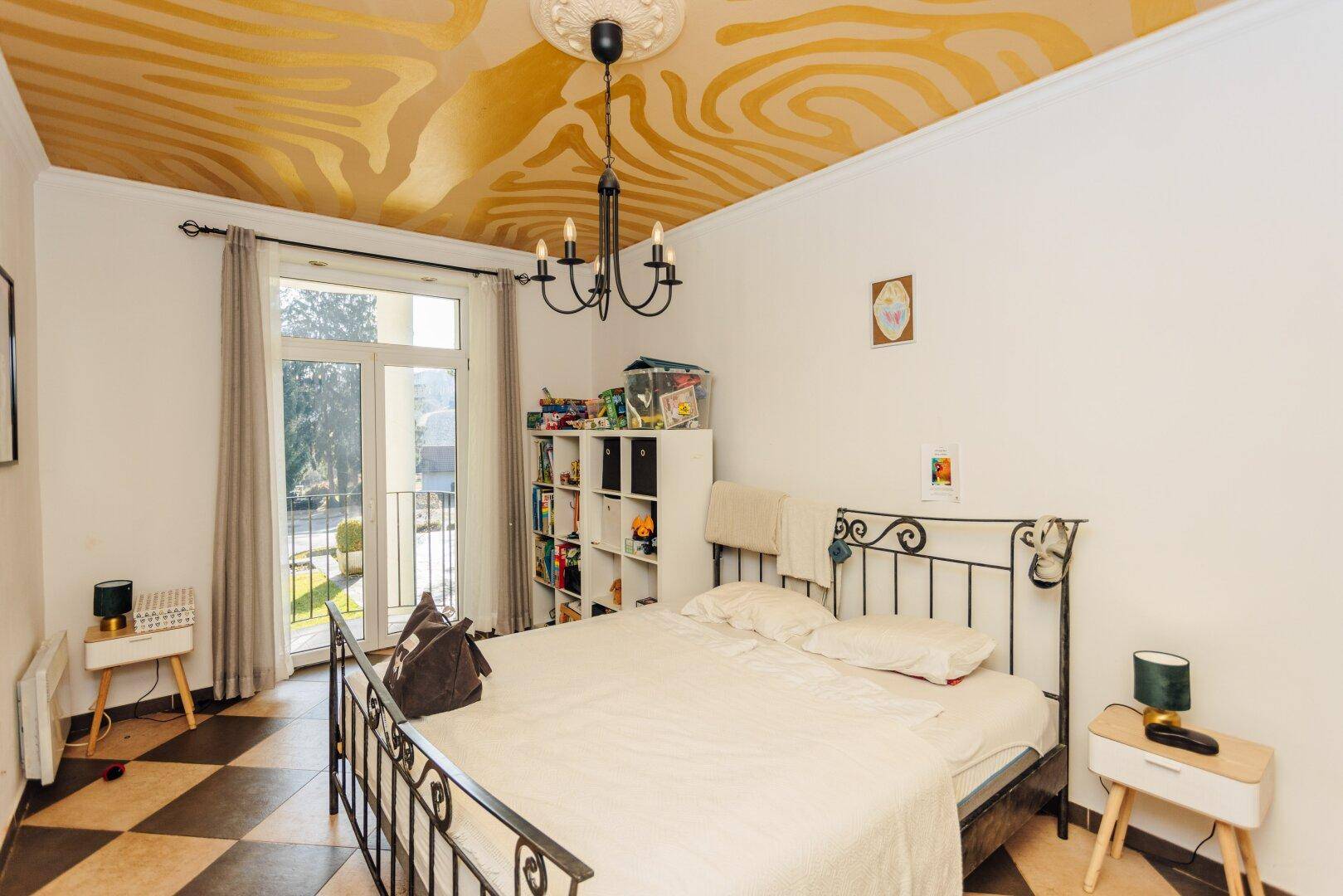 Schlafzimmer mit Metallbettgestell, gemusterter Decke und Zugang zum Außenbereich.
