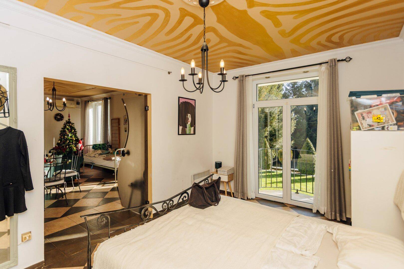 Helles Schlafzimmer mit Metallbett, gemusterter Decke und Blick in den Wohnbereich.