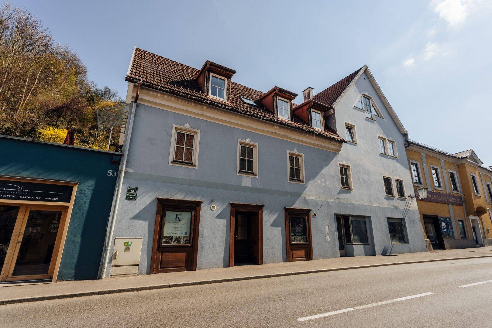 Ansicht des historischen Gebäudes mit hellblauer Fassade, mehreren Fenstern und rotem Ziegeldach.