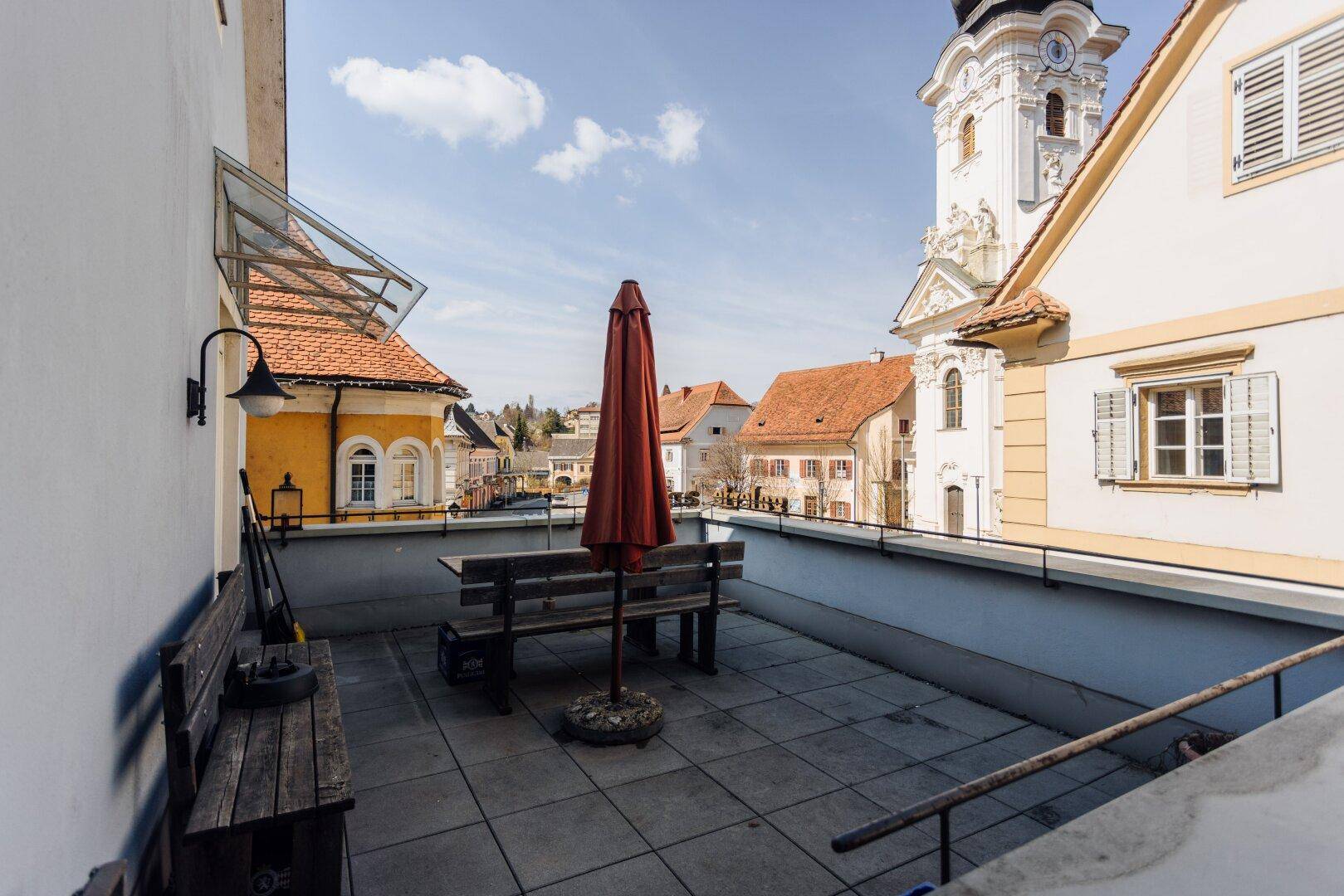 Gemütliche Terrasse mit Holzbänken und Sonnenschirm, bietet einen schönen Blick auf den Kirchturm.