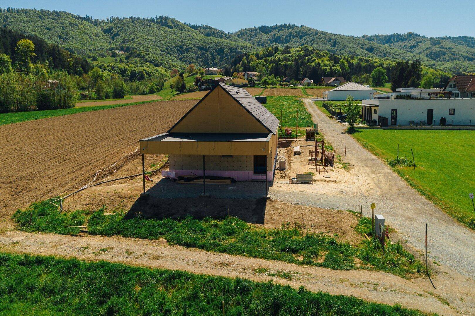 Luftaufnahme eines Neubaus in Holzbauweise mit Satteldach, umgeben von Feldern und grünen Hügeln.