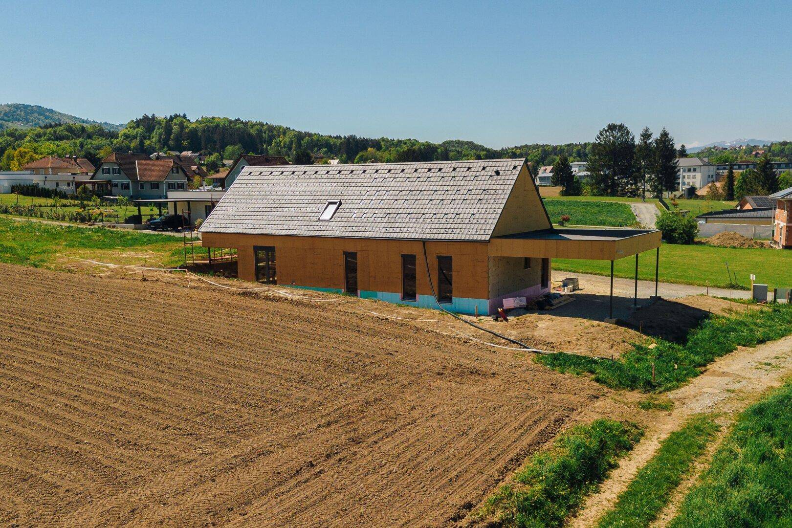 Seitenansicht eines Neubaus mit Satteldach und Dachfenster, umgeben von Ackerland und ländlicher Bebauung.