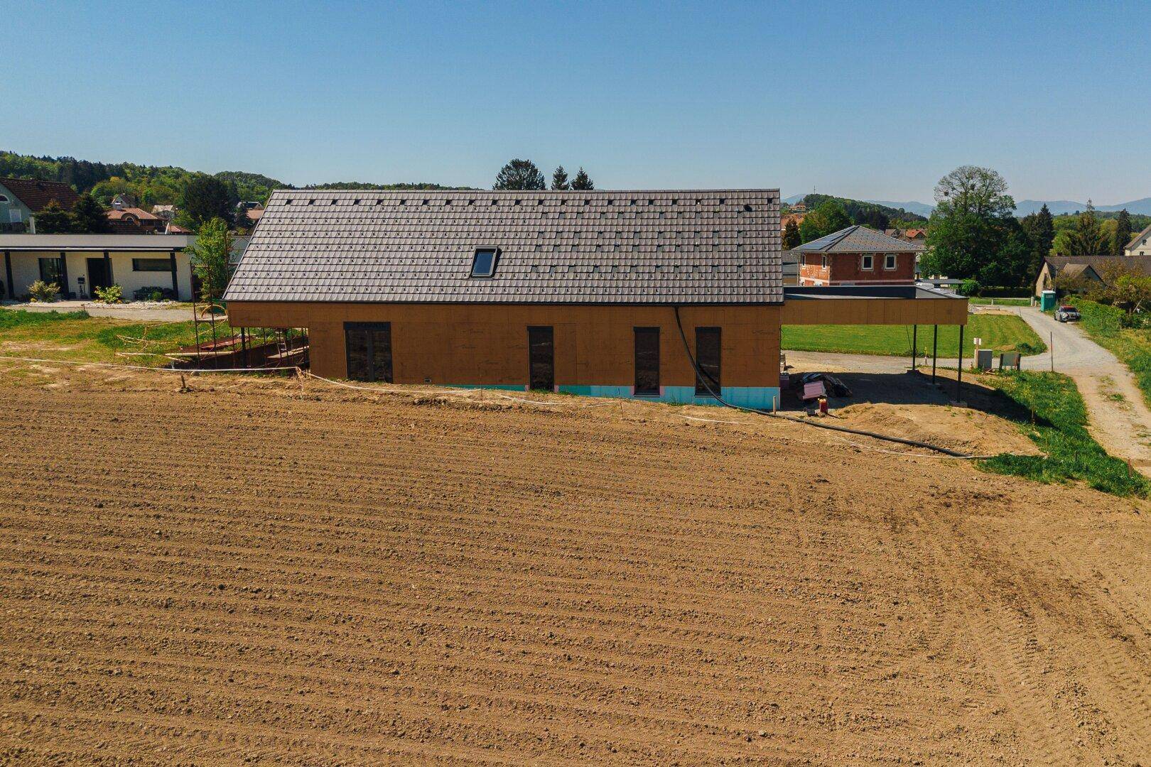 Frontale Luftansicht des Neubaus mit Satteldach und Dachfenster, umgeben von Feldern und Natur.