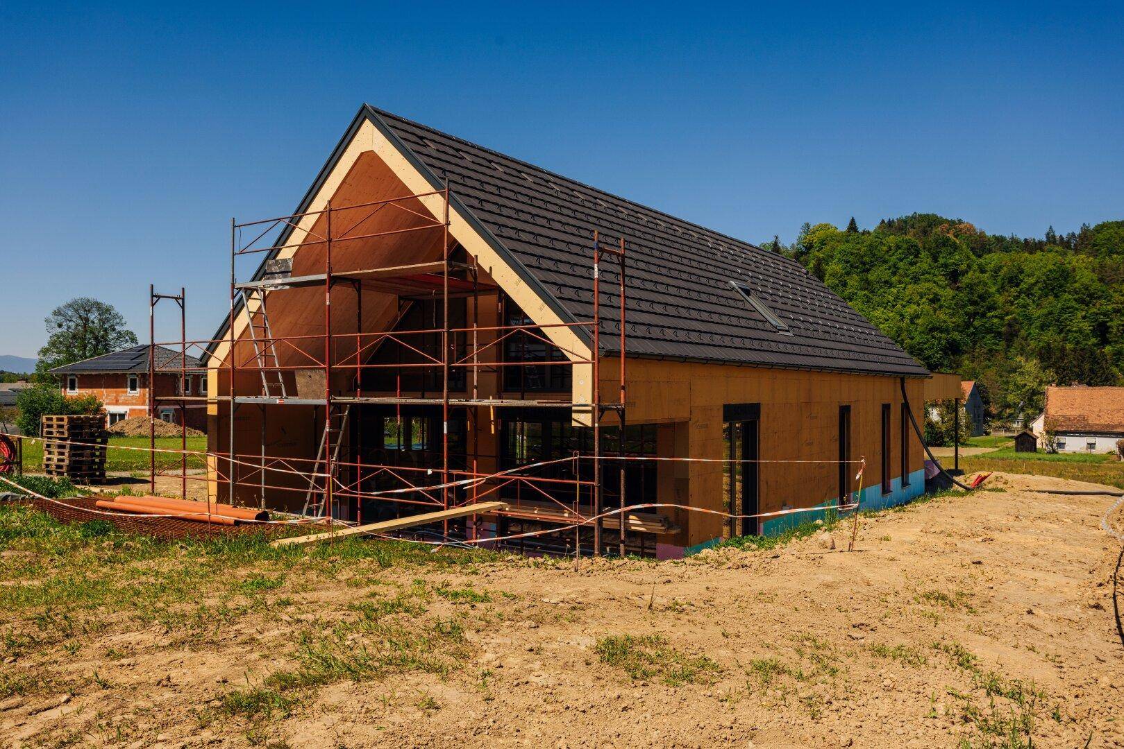Neubau in Holzbauweise mit Gerüst und sichtbarer Dachkonstruktion, umgeben von grünen Hügeln.