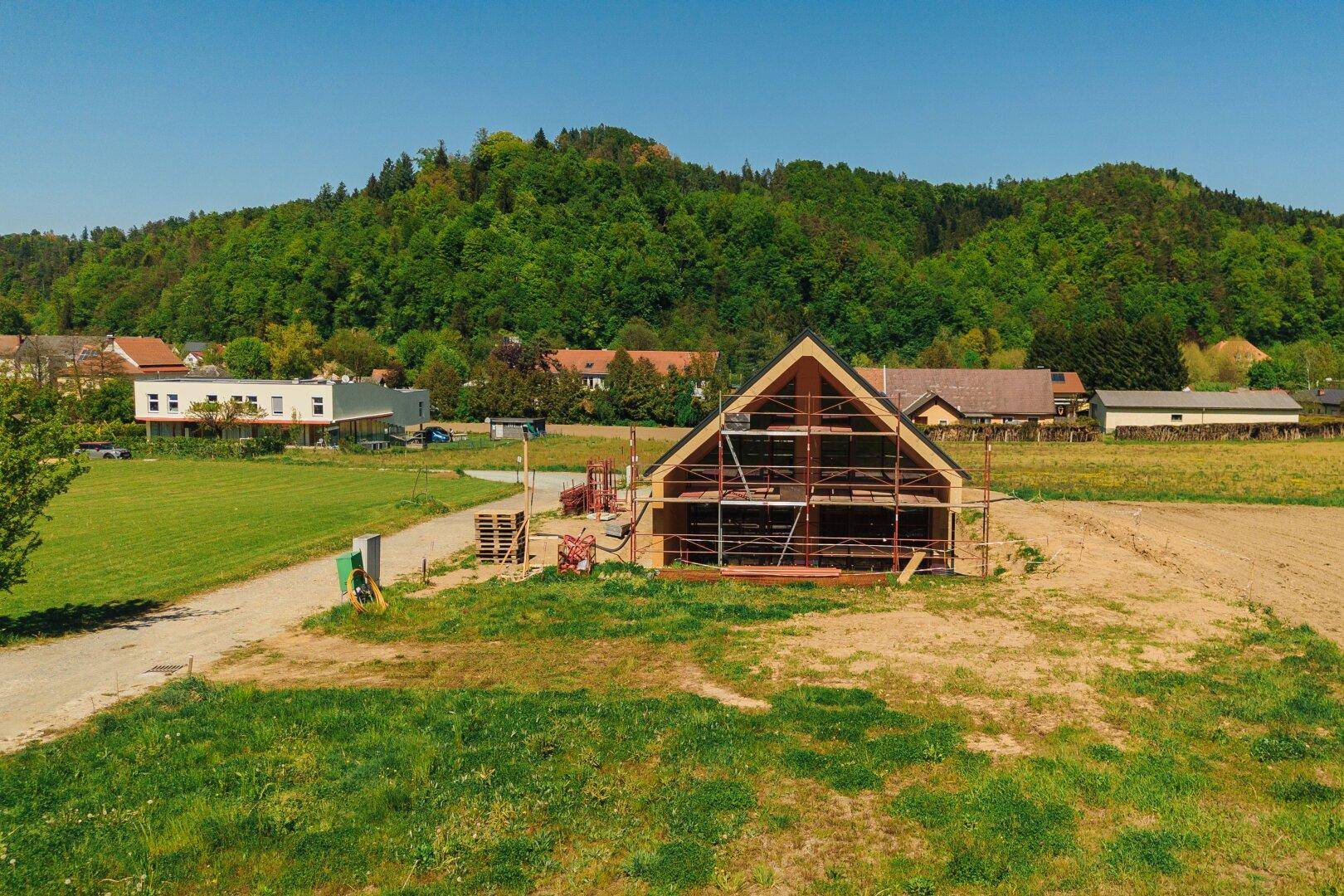Weitläufige Ansicht des Neubaus mit Gerüst in ländlicher Umgebung, umgeben von Wiesen und Wald.