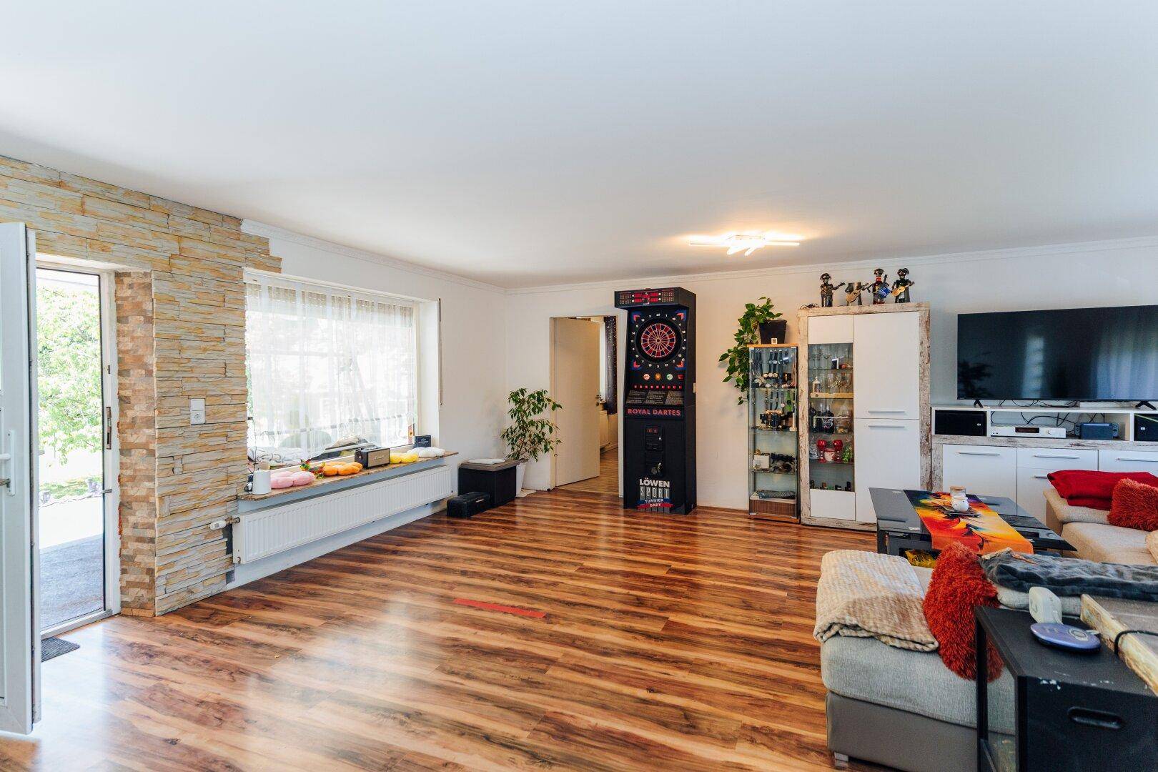 Helles Wohnzimmer mit Laminatboden, großem Sofa, Fernseher und einem Dartspiel.