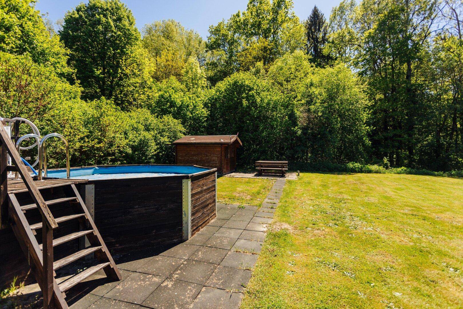 Aufstellpool mit Holztreppe, umgeben von Rasen, einem kleinen Schuppen und einer Sitzbank.