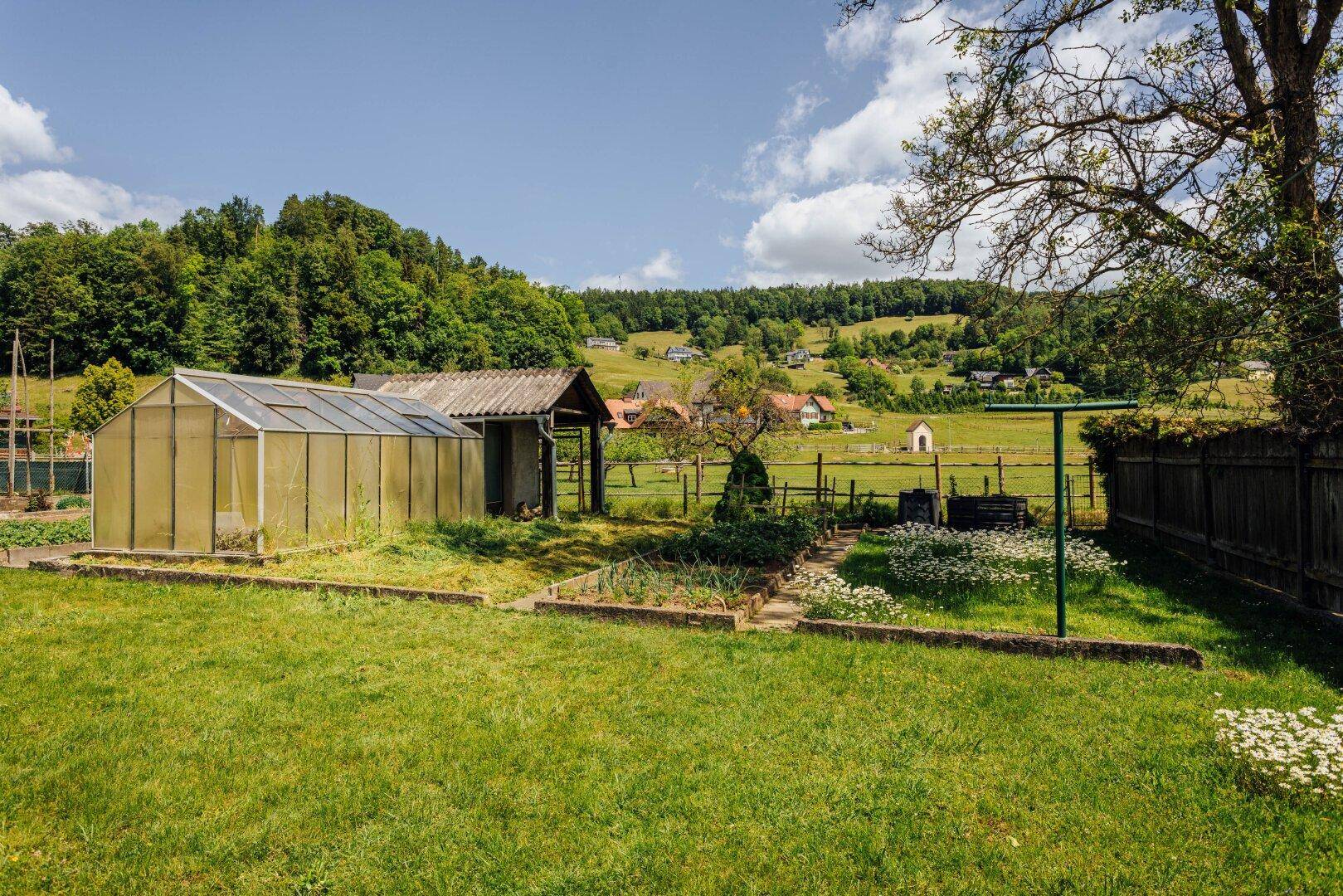 Garten mit Gewächshaus, kleinem Schuppen und angelegten Beeten vor hügeliger Kulisse.