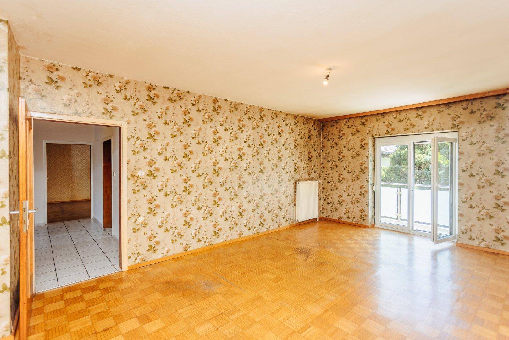 Leeres Wohnzimmer mit floraler Tapete, Parkettboden und Zugang zum Balkon.