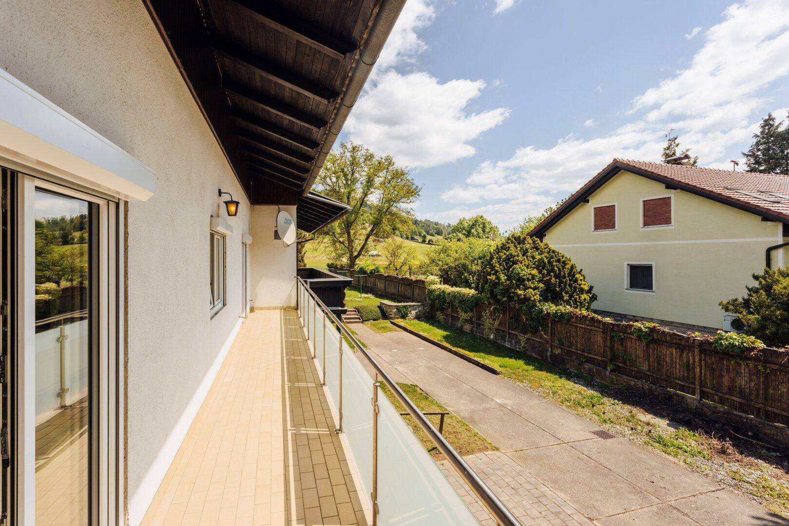 Langer Balkon mit Glasgeländer und Fliesenboden, mit Blick auf die Einfahrt und Grünflächen.