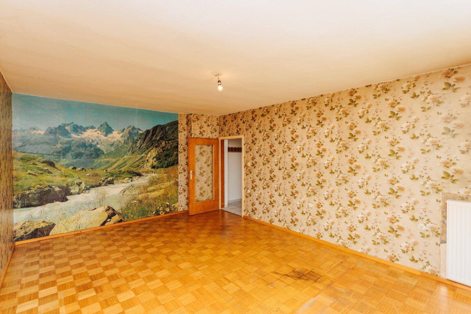 Leerer Raum mit floraler Tapete, Parkettboden und einer beeindruckenden Berglandschaft als Wandbild.