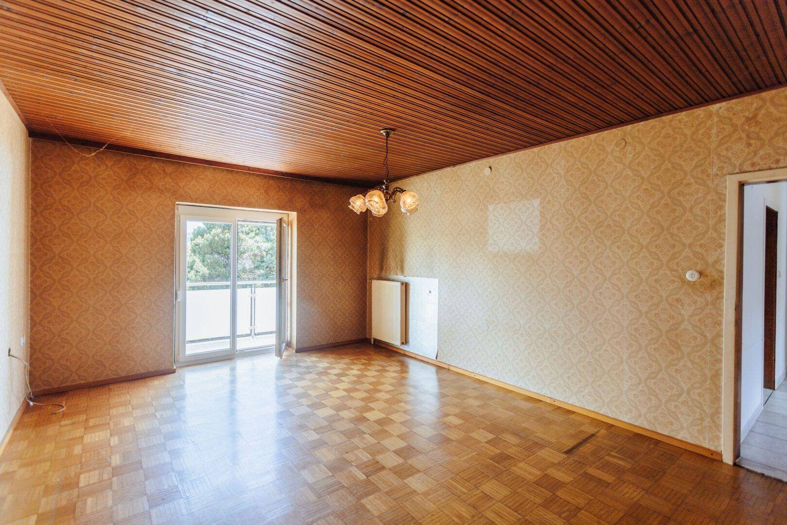 Leerer Raum mit gemusterter Tapete, Holzdecke, Parkettboden und Balkonzugang.