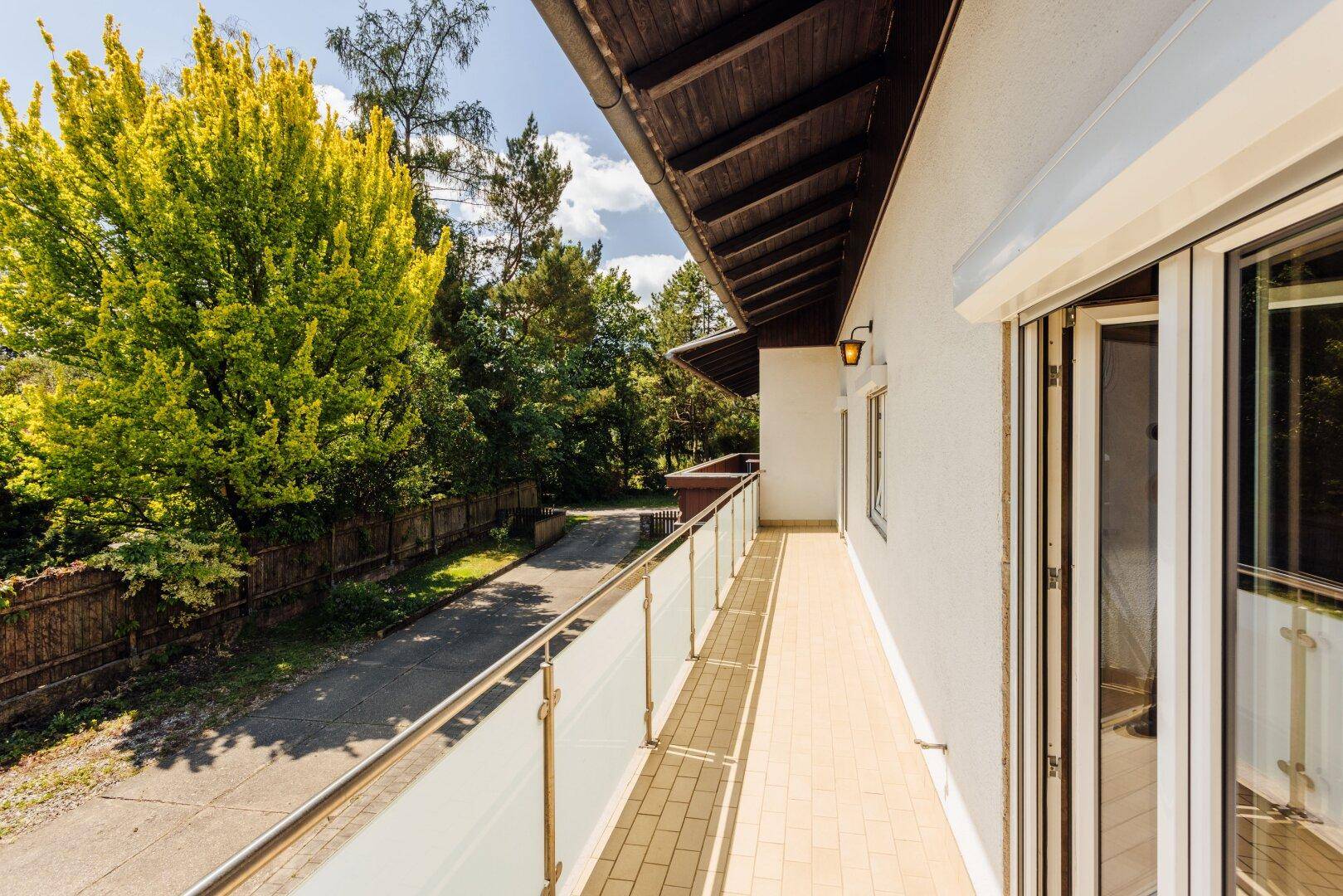 Langer Balkon mit Glasgeländer und Fliesenboden, mit Blick auf Bäume und die Einfahrt.