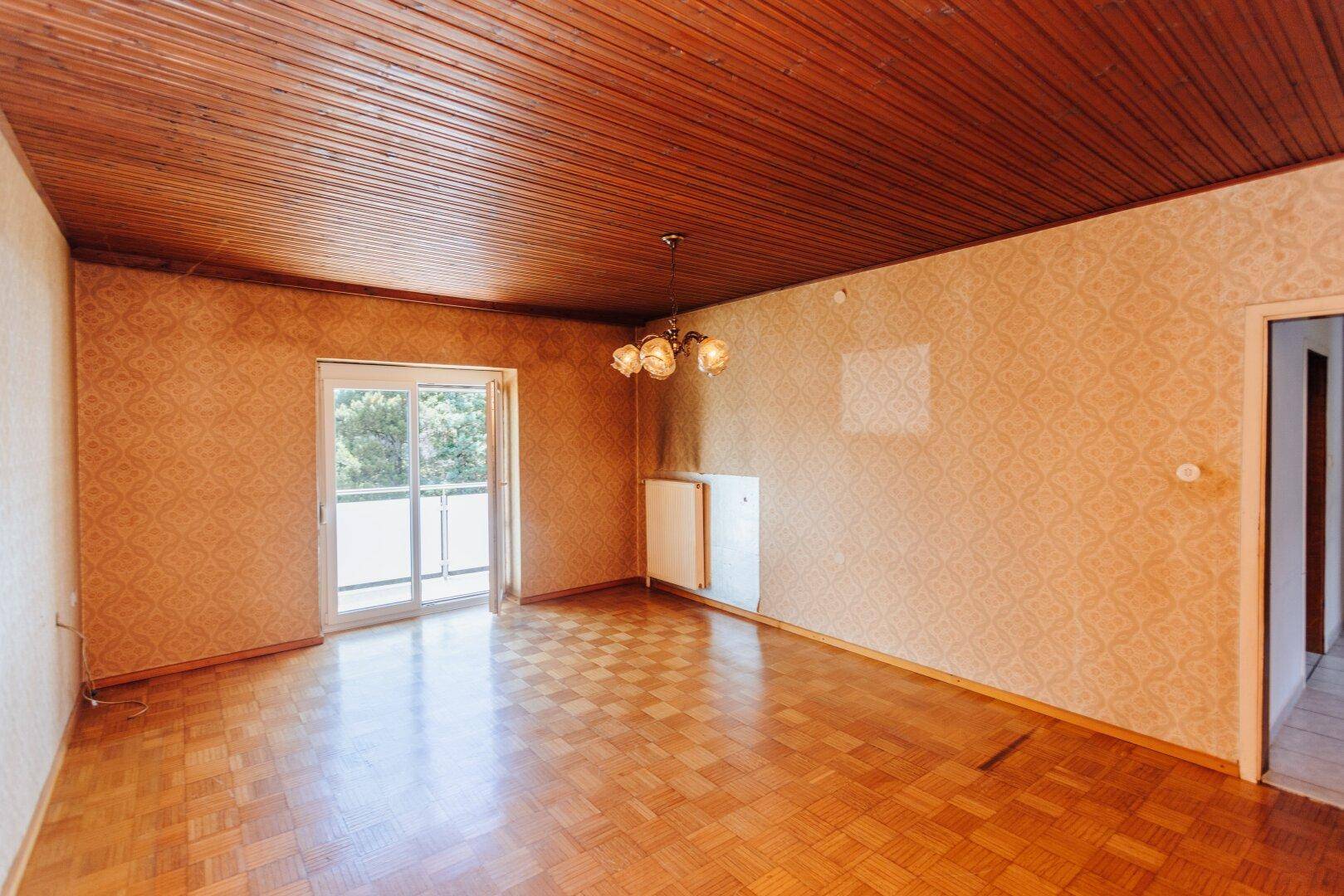 Geräumiges Wohnzimmer mit Parkettboden, Holzdecke und Zugang zu einem Balkon, renovierungsbedürftig.