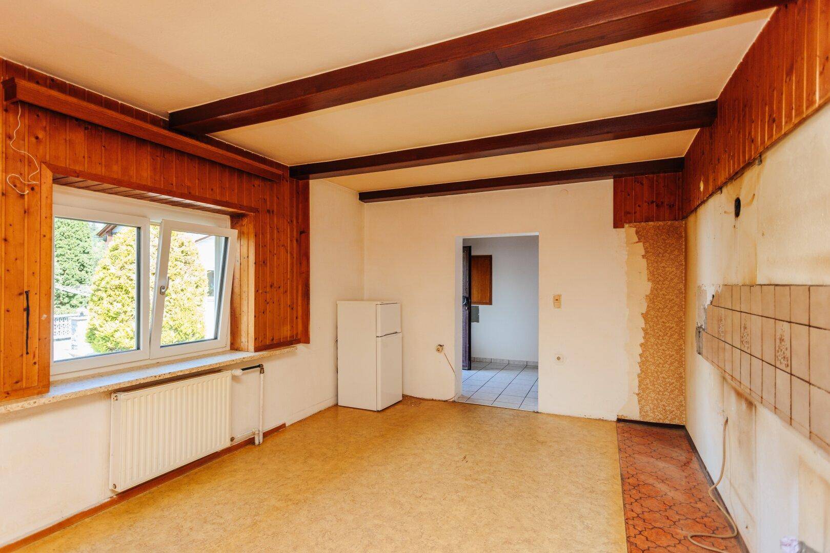 Renovierungsbedürftige Küche mit Holzvertäfelung, sichtbaren Balken und Fenster, bereit zur Neugestaltung.