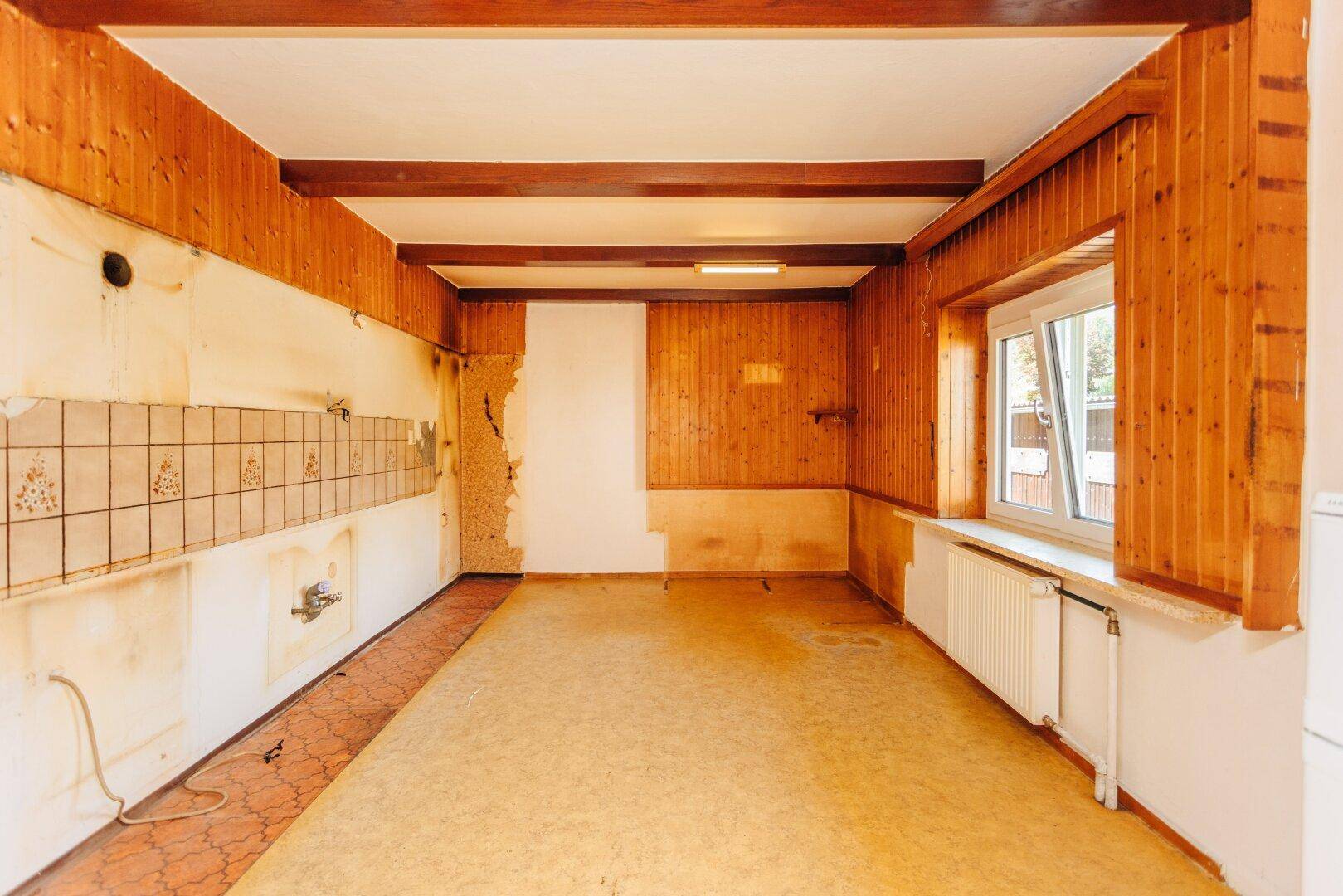 Leerer Küchenraum mit Holzvertäfelung, freigelegten Balken und Fenster, ideal für eine moderne Neugestaltung.