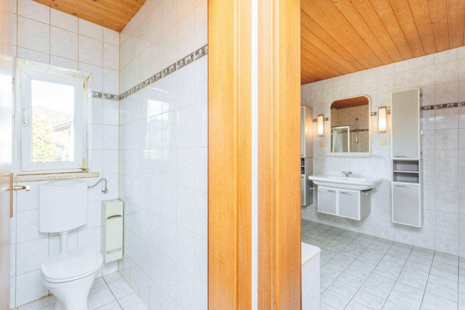 Badezimmer mit weißen Fliesen, Holzdecke, WC und Waschtisch, mit Fenster für Tageslicht.