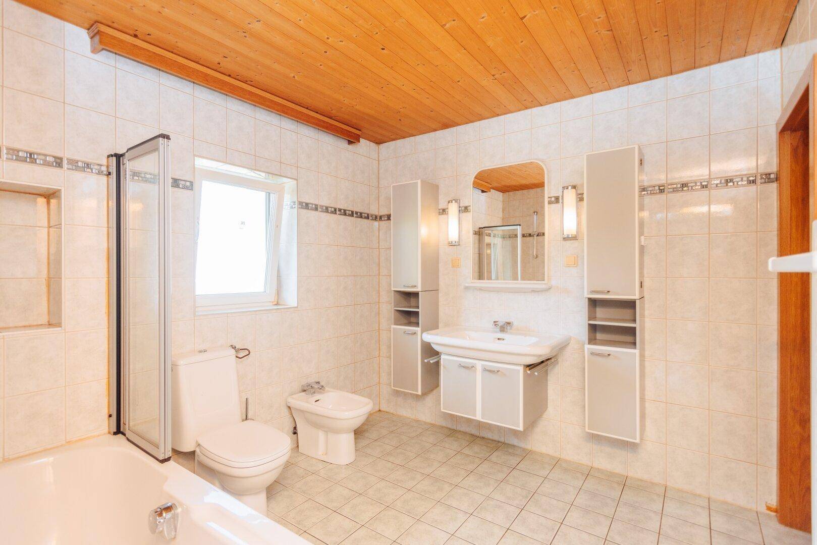 Geräumiges Badezimmer mit Badewanne, WC, Bidet und Waschtisch, unter einer Holzdecke.