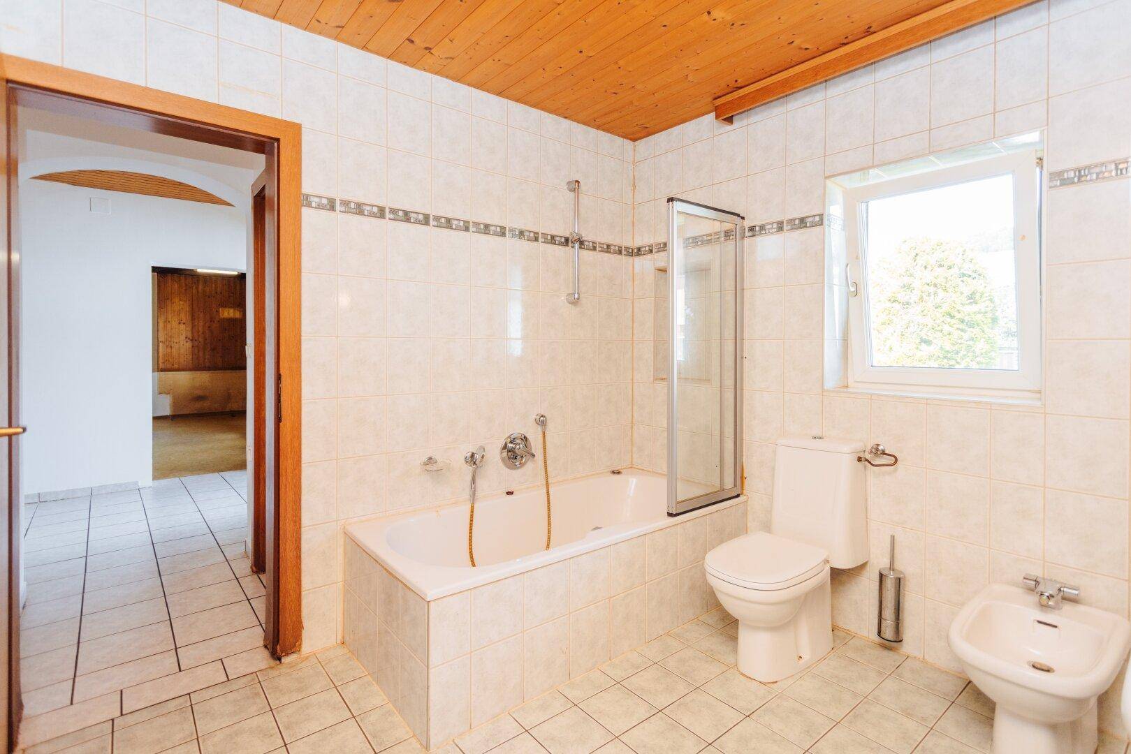 Badezimmer mit Badewanne, Duschabtrennung, WC und Bidet, unter einer rustikalen Holzdecke.