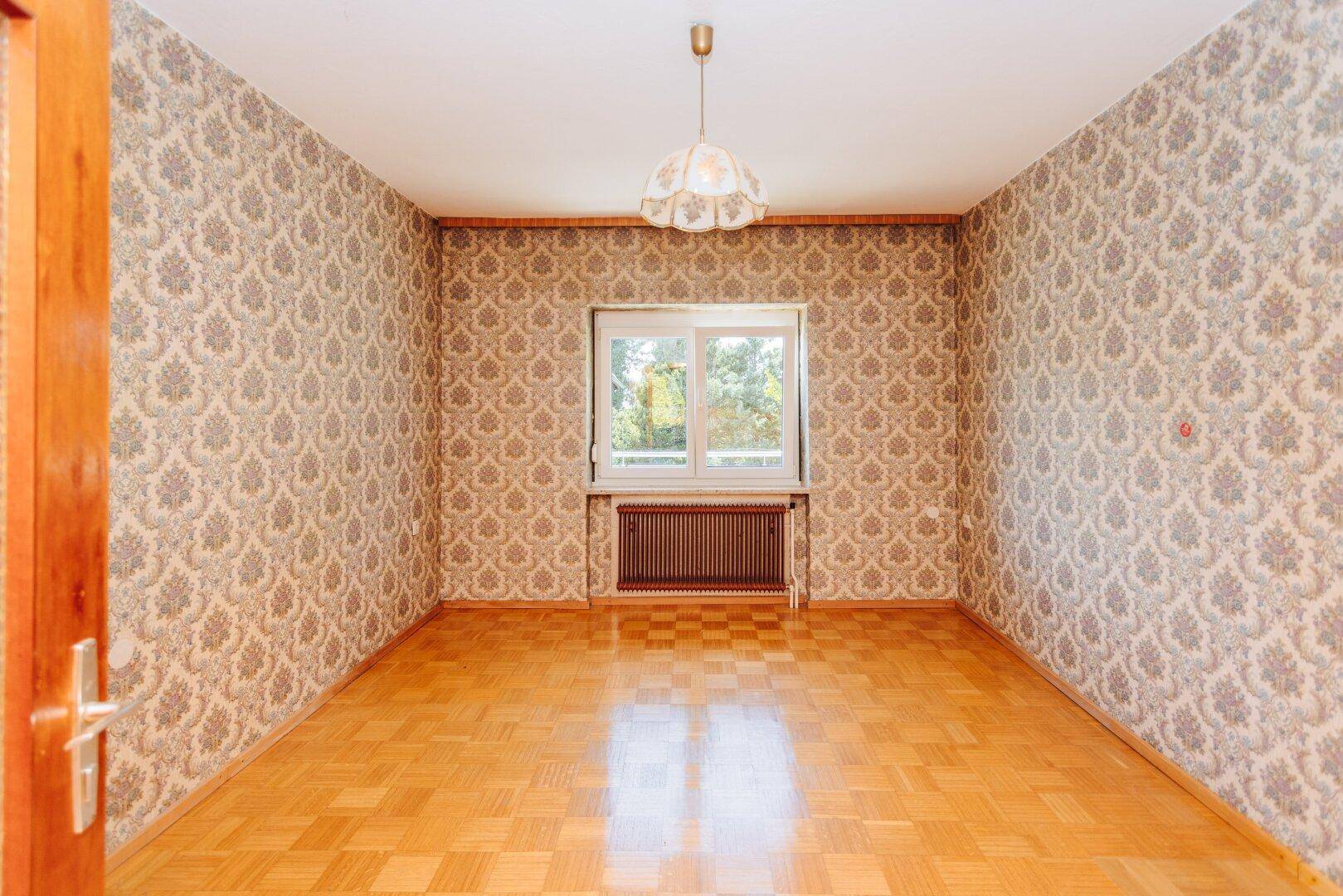 Schlafzimmer mit Parkettboden, gemusterter Tapete und Fenster, bereit für eine Modernisierung.