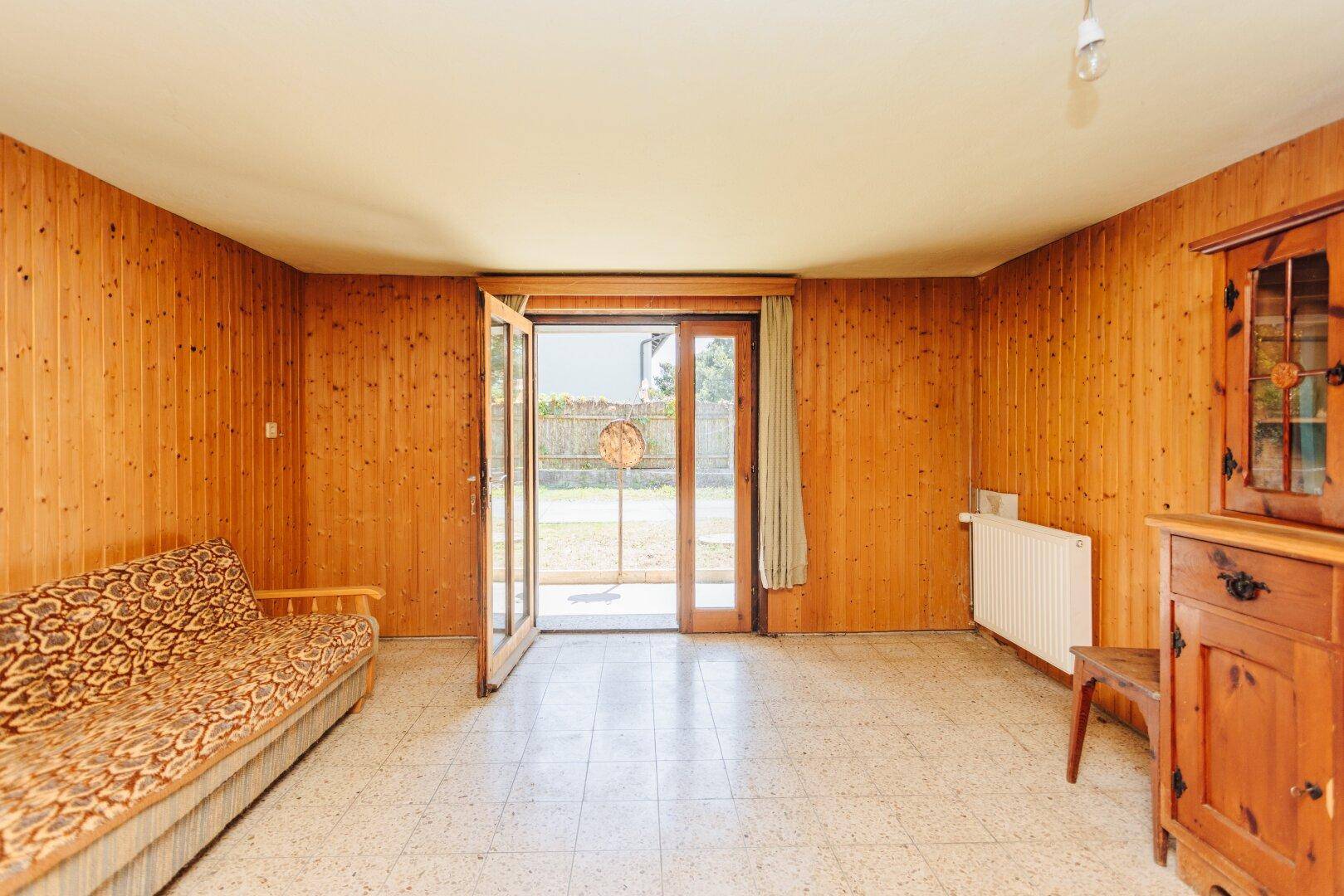 Heller Wohnraum mit Holzvertäfelung, Fliesenboden und großem Zugang zum Gartenbereich.
