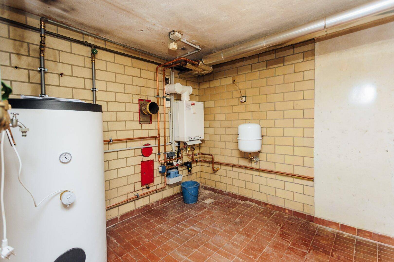 Heizungsraum im Keller mit Warmwasserspeicher, Heizkessel und sichtbaren Rohrleitungen.