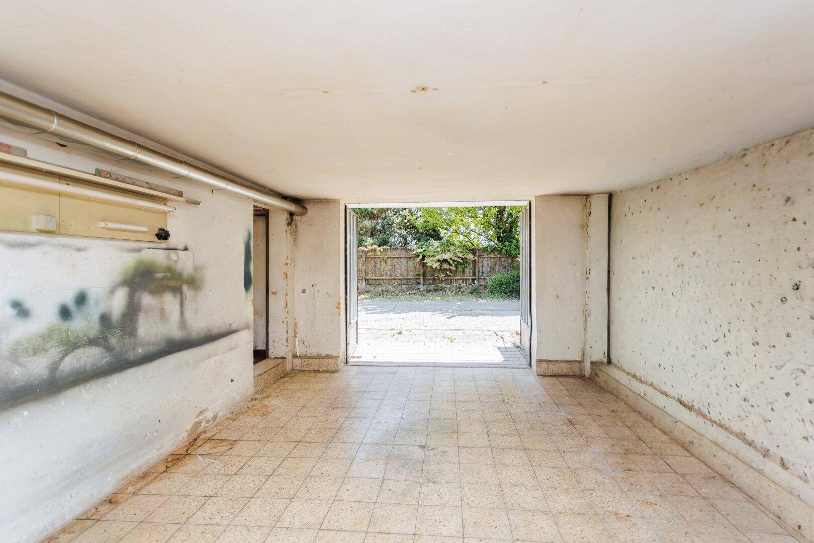 Leere Garage mit Fliesenboden und offenem Tor, bietet direkten Zugang zum Außenbereich.