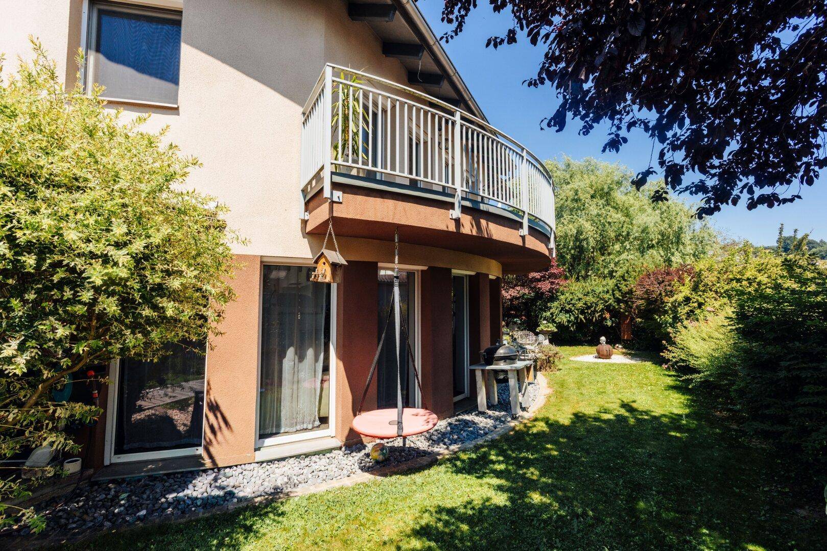 Detailansicht des Hauses mit geschwungenem Balkon und gepflegtem Gartenbereich im Sonnenschein.