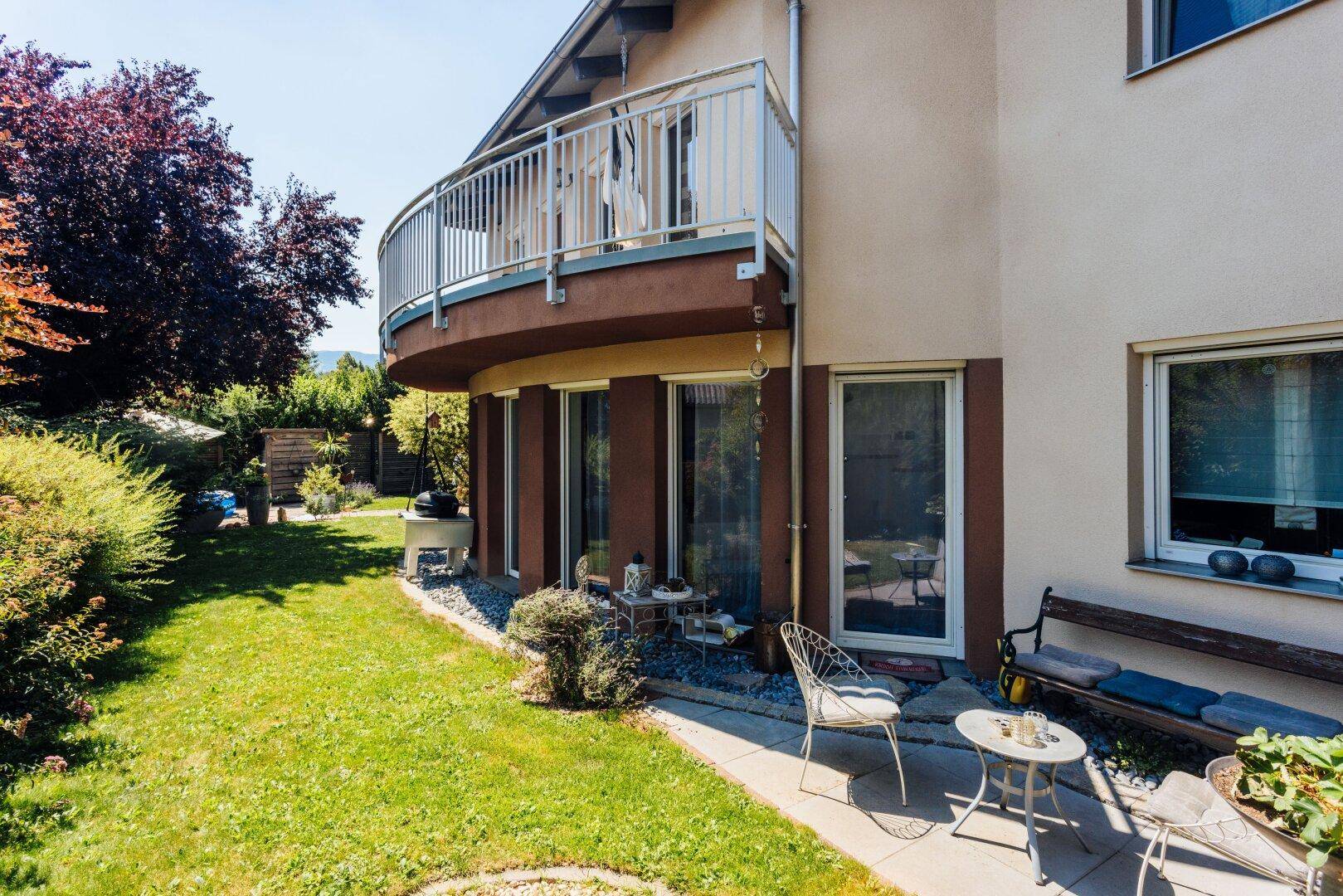 Hausfassade mit Balkon, Rasenfläche und einer kleinen, möblierten Sitzecke im Garten.