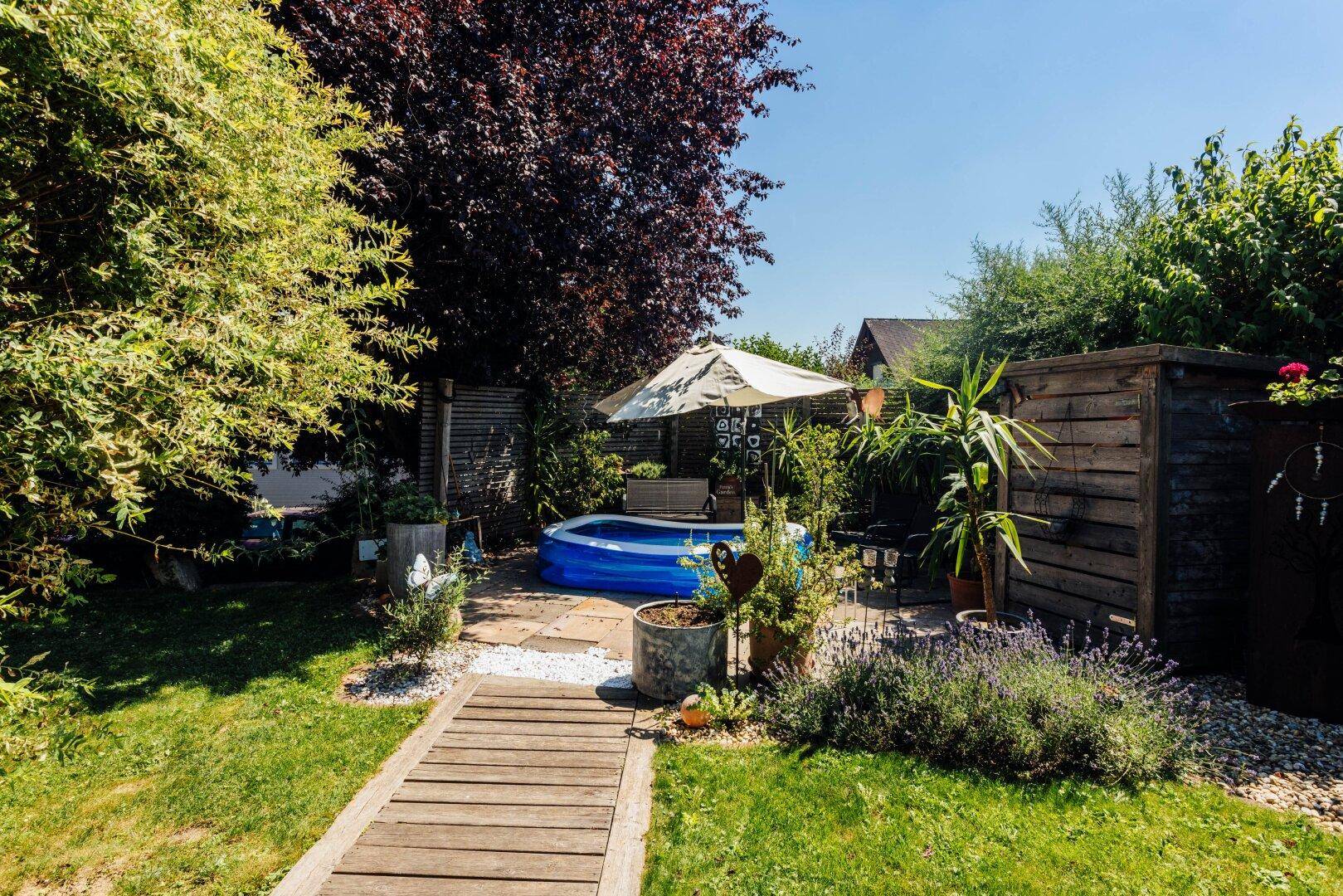 Gepflegter Garten mit Holzweg, kleinem Pool und Gartenhaus an einem sonnigen Tag.