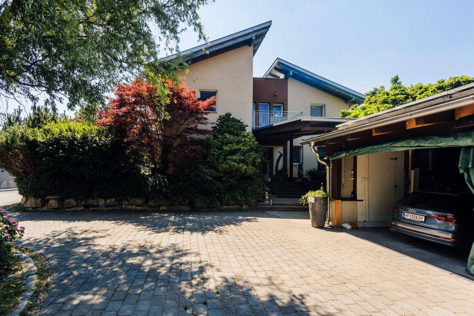 Vorderansicht des Hauses mit gepflasterter Einfahrt, Carport und üppiger Bepflanzung.