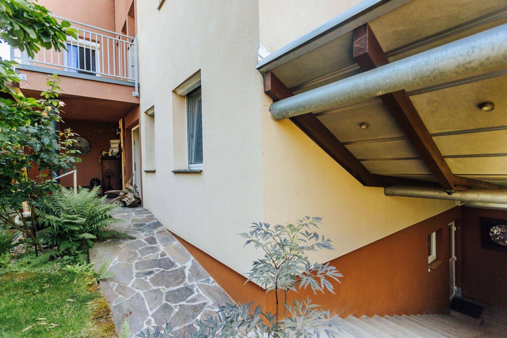 Seitenansicht des Hauses mit Steinweg, der zu einem unteren Bereich führt, umgeben von Grün.