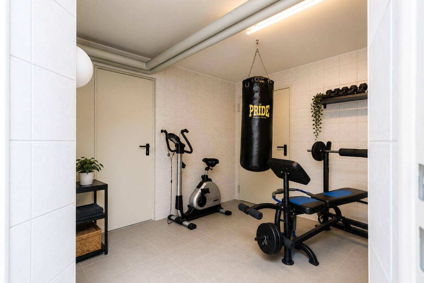 Heller Fitnessraum mit Trainingsgeräten, Boxsack und Fliesenboden für sportliche Aktivitäten.