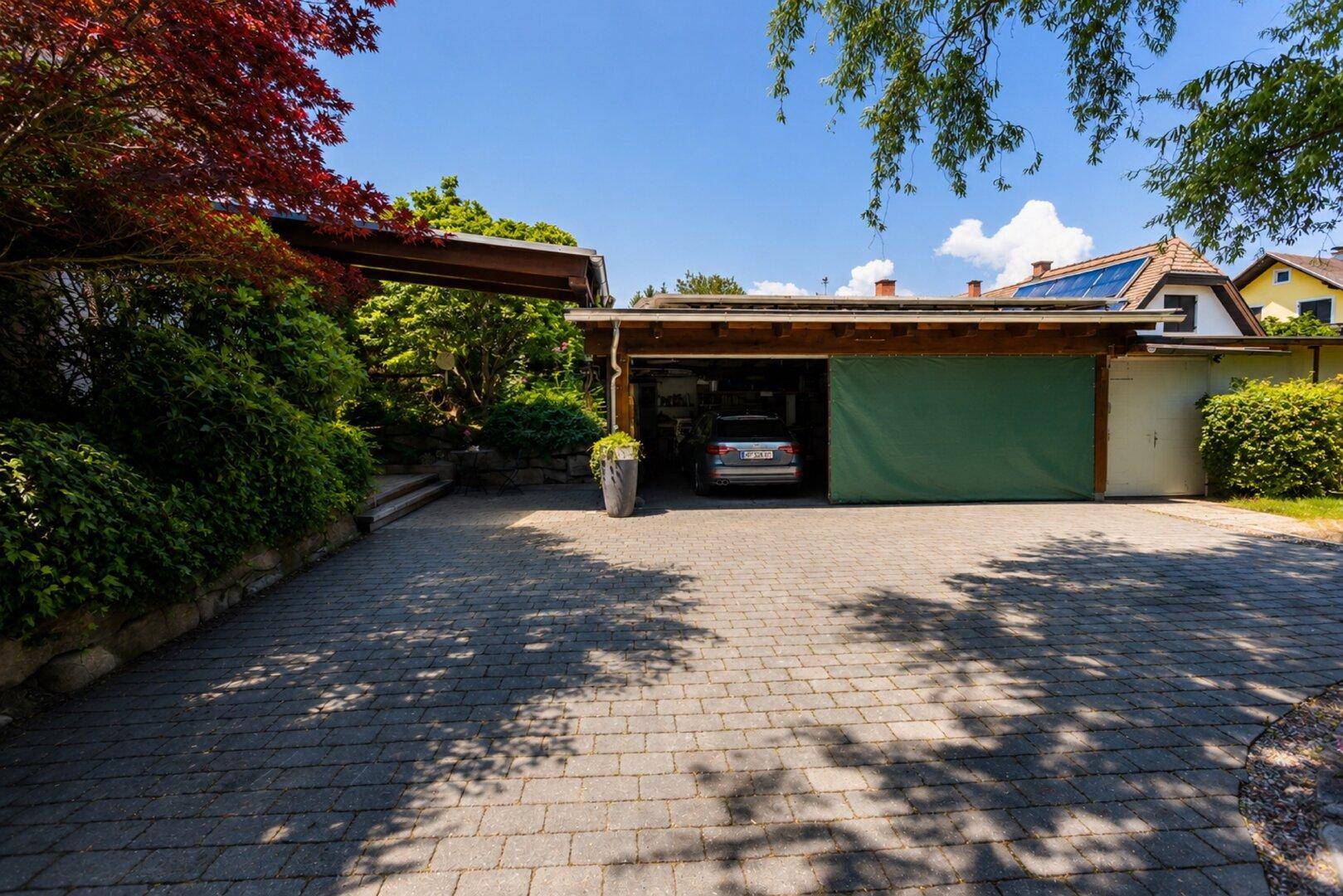 Gepflasterte Auffahrt führt zu einer Doppelgarage mit Carport und üppiger Bepflanzung.