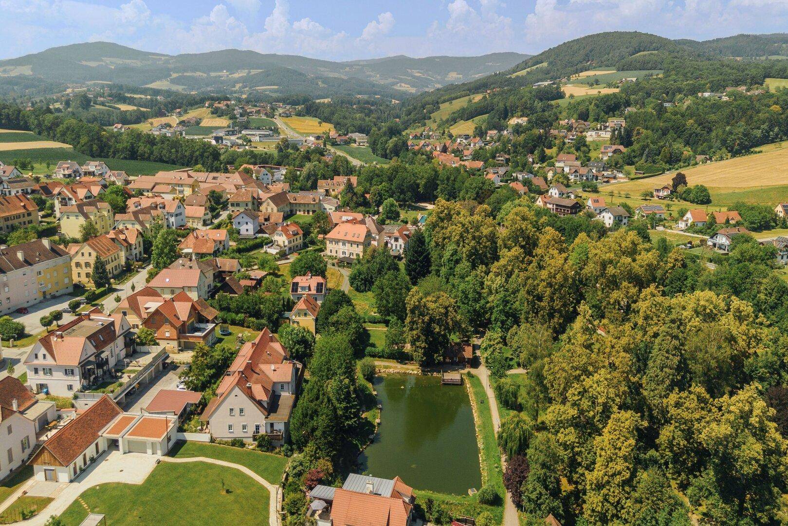 Malerische Luftaufnahme eines Dorfes mit Häusern, viel Grün und einem kleinen Teich in der Landschaft.