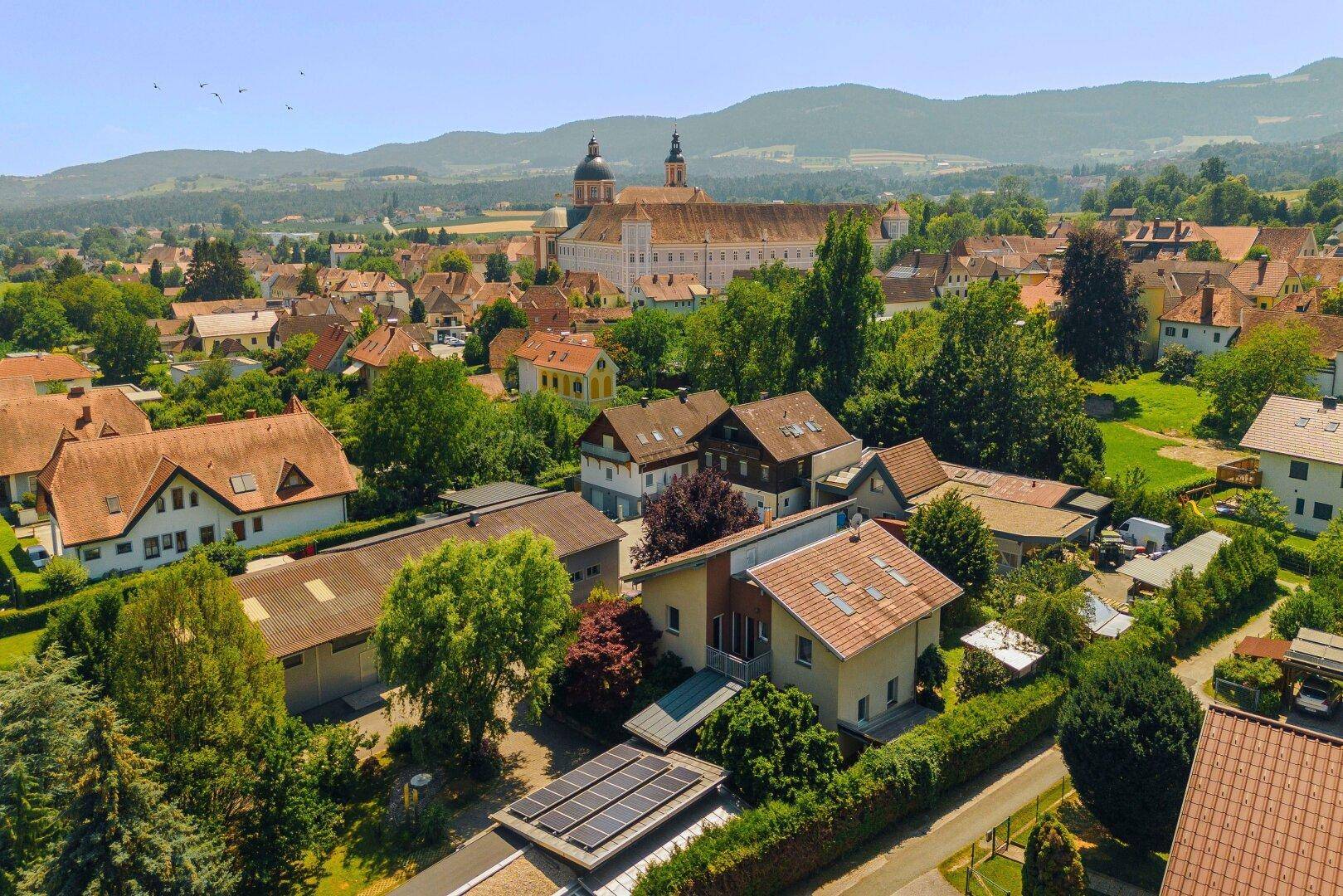 Weitläufige Luftaufnahme eines Dorfes mit Wohngebäuden, viel Grün und einem großen historischen Gebäude im Hintergrund.