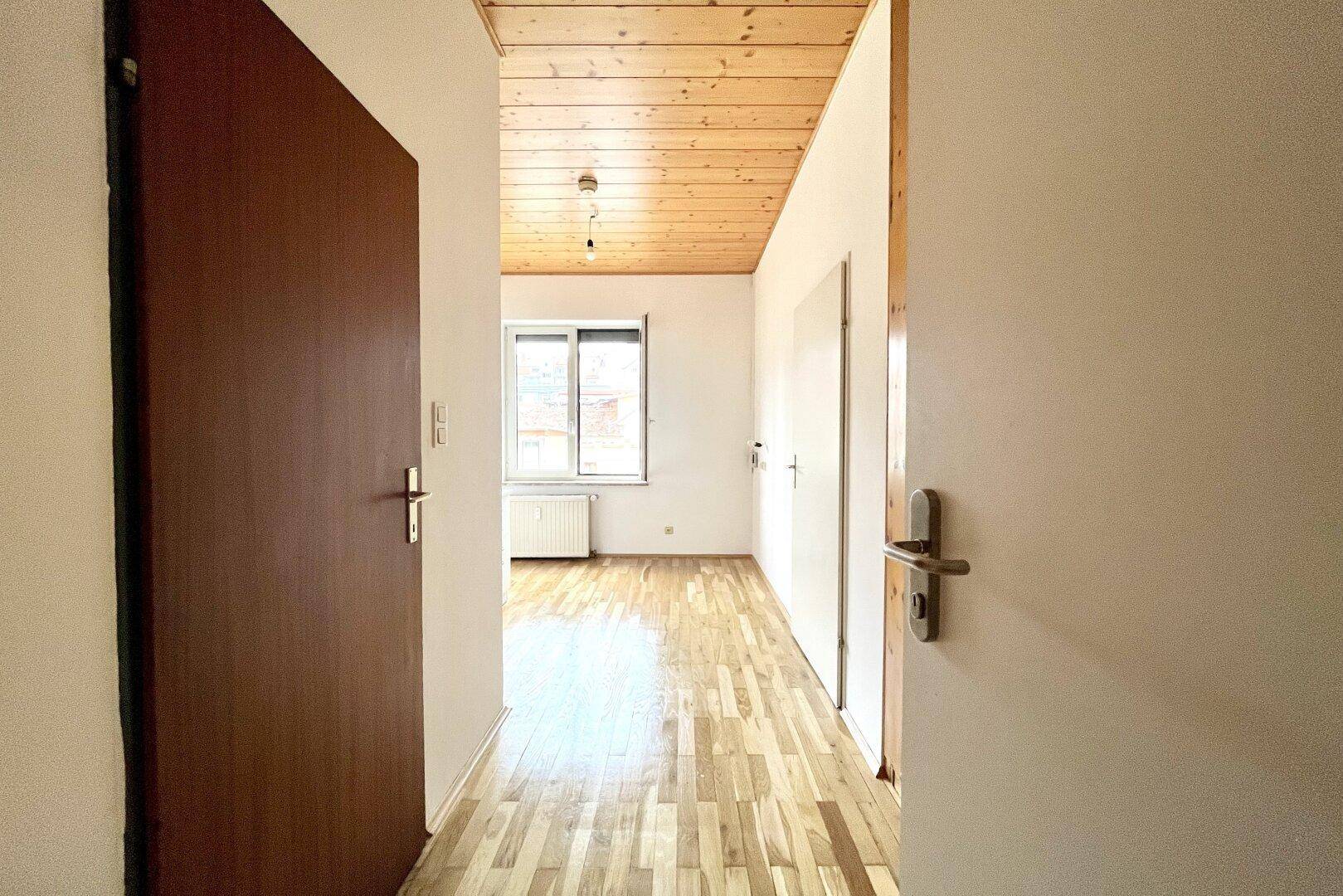 Heller Flur mit Holzbalkendecke und Parkettboden, der zu einem Zimmer mit Fenster führt.