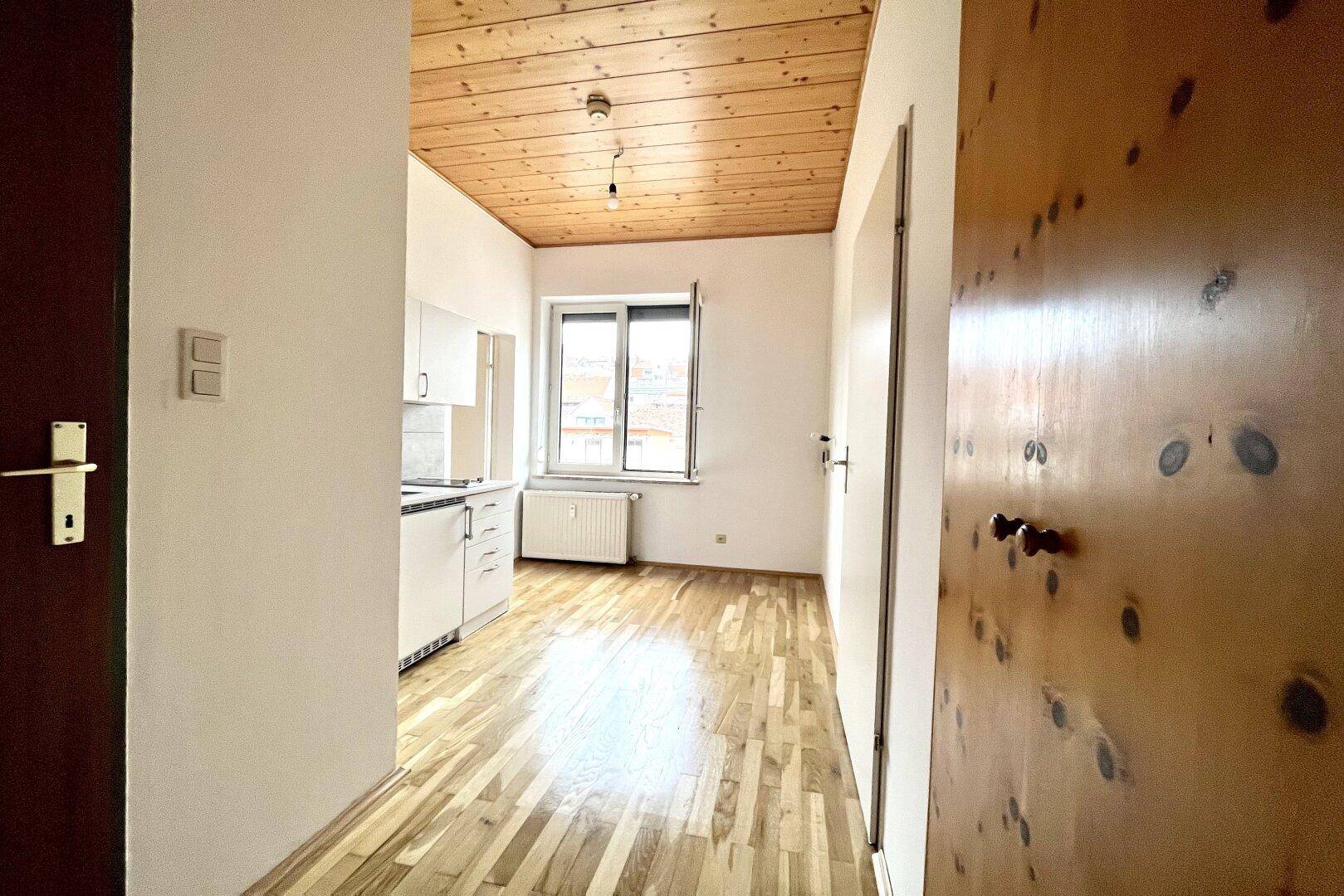 Kleiner Küchenbereich mit weißen Schränken und Holzbalkendecke, angrenzend an einen hellen Raum.