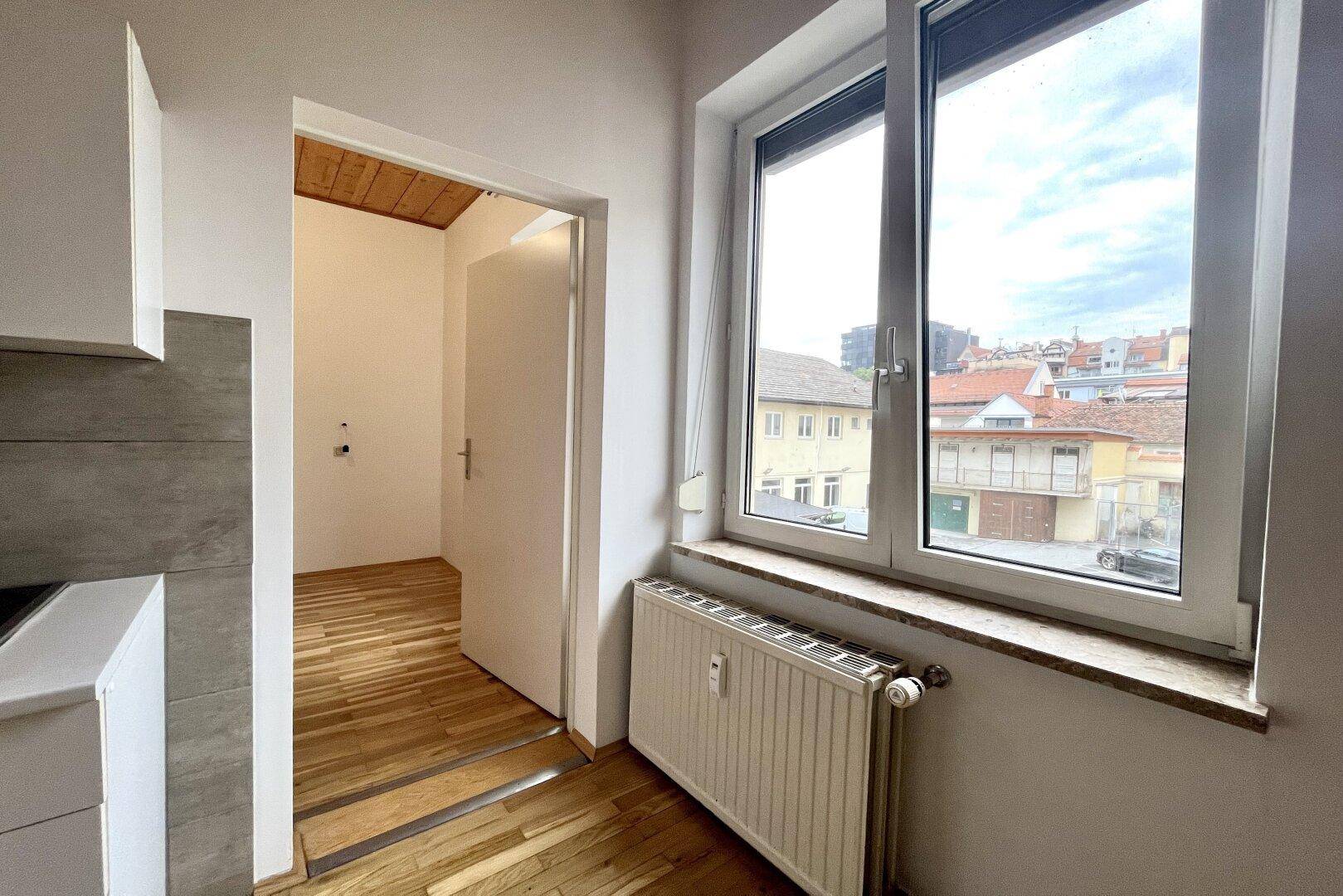 Blick aus dem Küchenbereich durch eine offene Tür, mit Heizkörper unter dem Fenster und Holzbalkendecke.